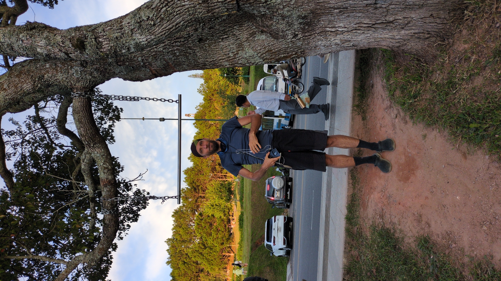 A full-body, vertical shot captures a man standing in front of a large tree in Da Lat, Vietnam. He is positioned centrally, facing the camera. He has a beard and is wearing a dark blue t-shirt, black shorts, black socks, and dark colored slip-on shoes. A black scarf is worn around his head. He is holding a phone in his right hand and headphones around his neck.
Behind the man, a street is visible with cars parked along it. A white SUV is on the left, followed by a darker SUV. A white van is further down the street, and a person is walking past it, carrying a bag. A motorcycle with a rider is also seen on the street.
In the background, a hill covered with pine trees slopes upwards, with some buildings or structures visible on the crest. The sky is partly cloudy. The man is standing on a reddish-brown dirt path with patches of grass.
The scene appears to be set in a park or a scenic area near a road, likely a popular tourist spot in Da Lat, Vietnam, given the presence of pine trees and the general ambiance. The lighting suggests it might be late afternoon or early evening.