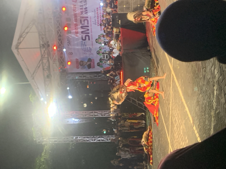 Two people are performing a traditional dance in front of a stage in Kecamatan Batu, Indonesia. One dancer, wearing a yellow and red costume with a large feathered headdress, is in mid-air, kicking their right leg forward. The other dancer, wearing a similar costume with a white headdress, is kneeling on the ground, facing the audience. Both performers have their faces obscured by their masks. The stage is decorated with red and white banners, a large banner displays an inscription in Indonesian. There are many people seated in the audience, most with their backs turned to the camera, Some members of the audience are holding up cellphones, possibly recording the performance. The performance is taking place outdoors on an asphalt or concrete surface, lit by overhead street lamps. It is night time and the lighting is dim. The location, based on the coordinates provided, is a street in the city of Batu, East Java, Indonesia.