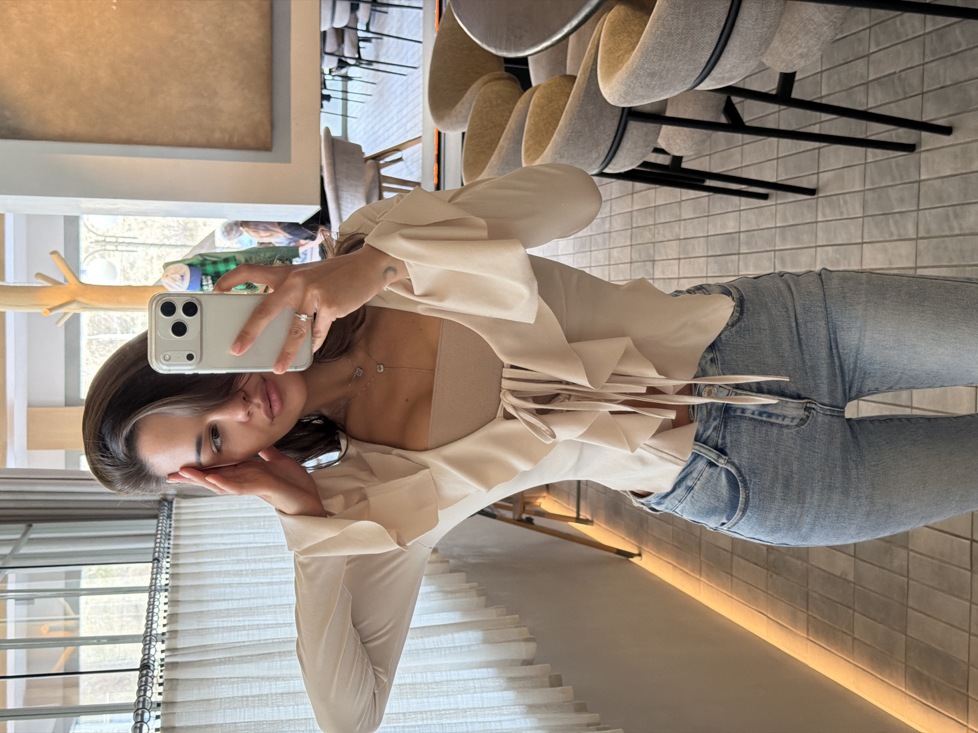 The image captures a woman taking a mirror selfie in a modern indoor setting, likely a cafe, restaurant, or lounge, in Krasnoyarsk, Russia. The woman, with dark brown hair, is dressed in a light cream or off-white ruffled blouse worn over a beige bandeau-style top, paired with light-wash blue jeans. She is adorned with a delicate necklace featuring two small pendants (one appearing clover-shaped, the other square) and a ring on her right ring finger.
She holds a light-colored smartphone, consistent with an iPhone 15 Pro Max in a clear case, with her right hand to capture her reflection. Her left hand is gently touching the side of her face, and she looks directly into the camera with a calm, composed expression.
The background features contemporary interior design elements. To her right, several light grey upholstered chairs with black metal frames are visible, arranged around tables. The floor is covered in light grey rectangular tiles. To her left, a large window with white horizontal blinds offers a glimpse of outdoor greenery and trees, suggesting daylight. A unique, light wooden, branch-like coat rack is partially visible near the window. Warm, indirect lighting softly illuminates parts of the wall. The overall ambiance is chic, clean, and minimalist. The image is oriented 90 degrees clockwise from standard portrait. No visible text is present within the frame.