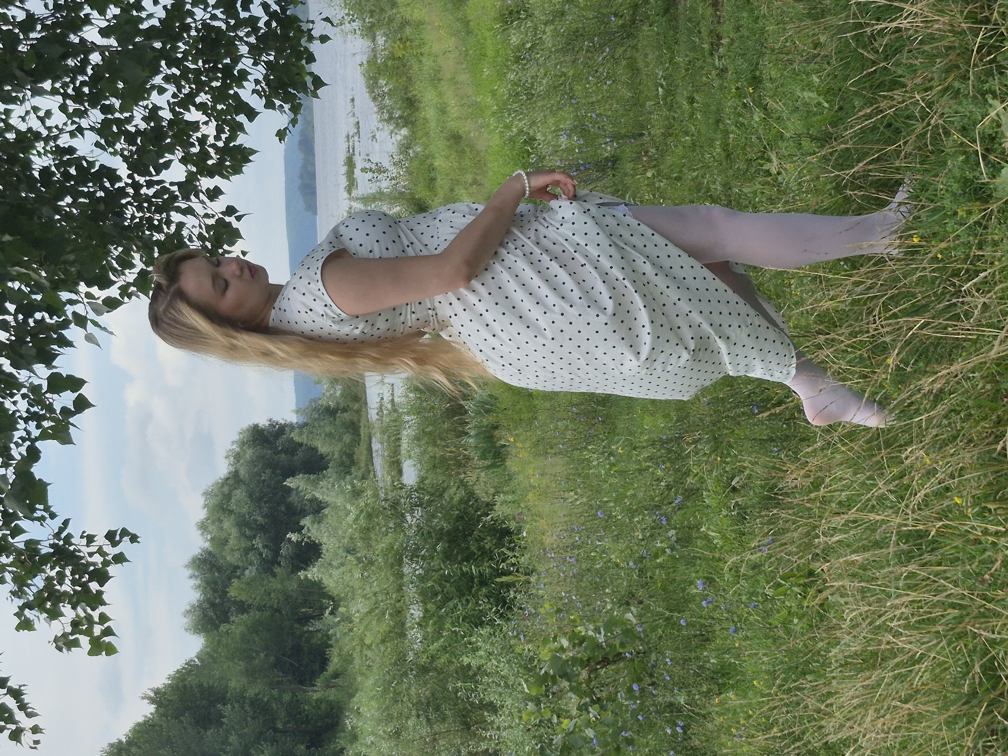 A young woman with long blonde hair stands in a grassy field near a body of water. She is wearing a white dress with black polka dots, white pantyhose, and white shoes. The woman is facing away from the camera, looking to the left. The background features trees and a cloudy sky. The scene appears to be in the city of Chaikovsky, Russia, given the geographical context often associated with this type of landscape. The environment is natural, with lush vegetation and a calm atmosphere.