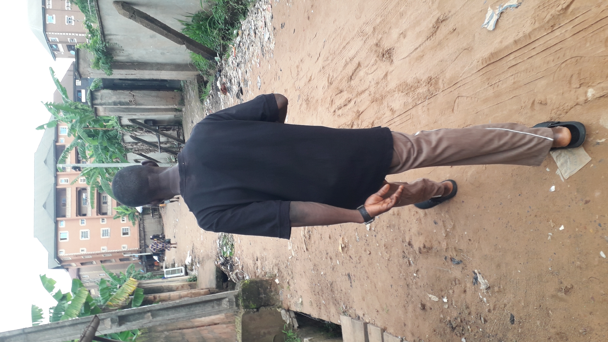 A full shot, taken from behind, captures a young Black male walking away from the camera down a dirt path. He is wearing a plain black t-shirt and light brown pants with a white stripe down the side of each leg. He has black sandals on his feet and a black watch on his left wrist. The path is flanked by concrete walls, some of which are weathered and cracked. Banana trees are visible behind the walls, and in the background, several multi-story apartment buildings can be seen, suggesting an urban environment. The ground is covered in loose dirt and scattered debris, including pieces of paper and plastic. The overall impression is a glimpse into a residential area in Nkpor, Nigeria, with elements of both ongoing construction and daily life.