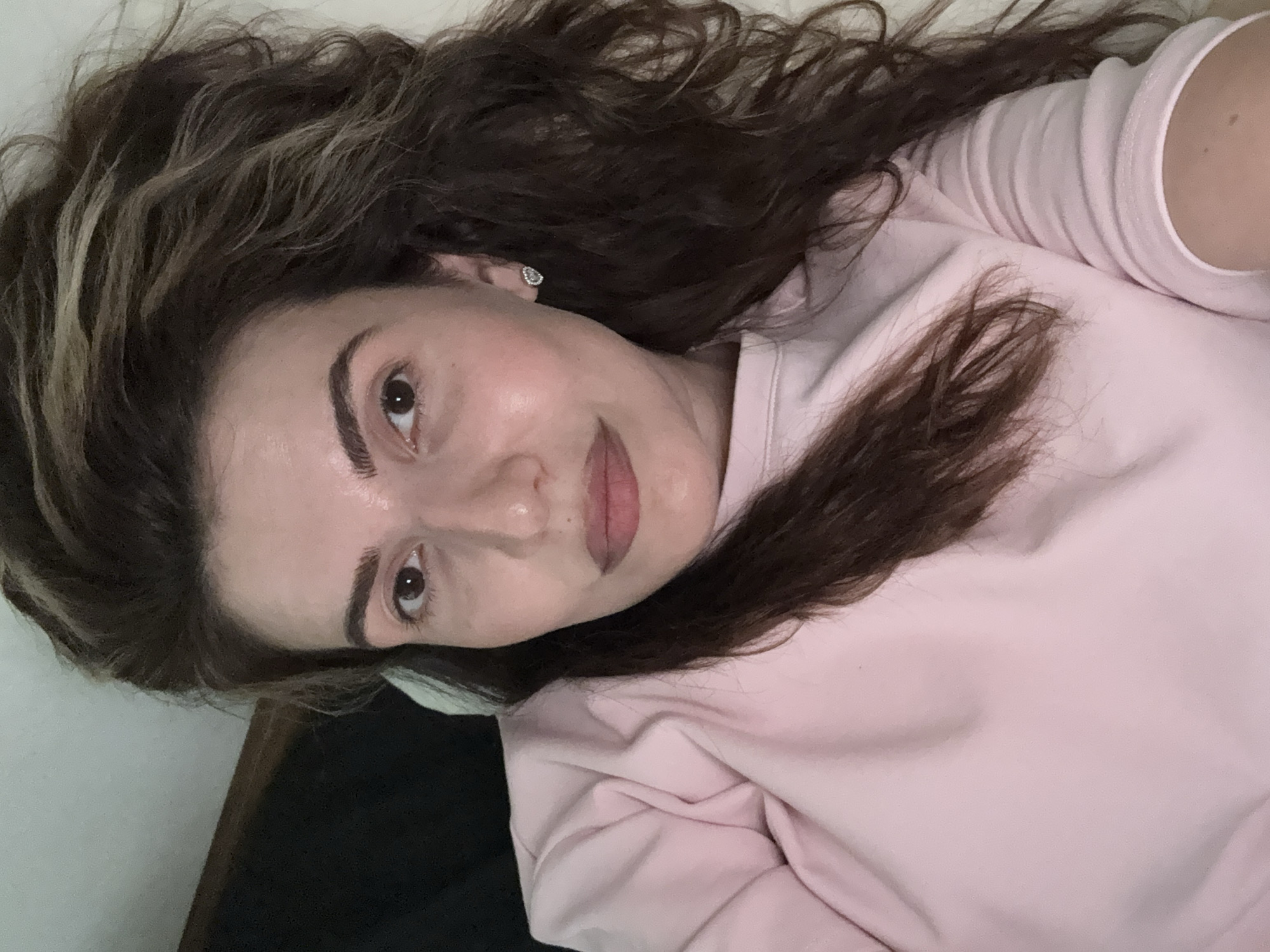 A medium shot captures a woman from the chest up, lying on her side, with her head resting against a dark, textured surface. She has dark brown hair with lighter streaks, styled in loose waves that fall across her shoulders and face. Her skin appears smooth and lightly made-up, with a noticeable sheen on her forehead and cheekbones, suggesting soft lighting. She is wearing a pink long-sleeved top. Her left eye is clearly visible, a deep brown, framed by dark eyebrows and subtle eye makeup. Her right eye is partially obscured by her hair. A small, heart-shaped earring with clear stones adorns her right ear. Her lips are softly closed, painted in a muted pinkish-brown shade. The background is a light, neutral color, likely a wall or bedding, with a darker area to the left, possibly a piece of furniture or shadow. The overall impression is one of casual relaxation.
