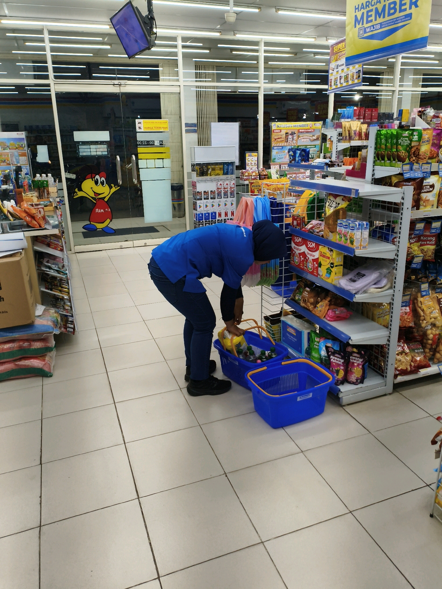 In a convenience store in Sumberejo Wetan, Indonesia, a person wearing a blue uniform and a black hijab is bending down to place items into a blue plastic shopping basket. The person is positioned in the middle aisle of the store, between shelves stocked with various products. A second, larger blue basket is on the floor nearby. The store's interior features white tiled flooring and fluorescent lighting. Prominent signage indicates membership benefits, including "HARGA TERMURAH MEMBER WAJIB UNTUK DAPAT HARGA KHUSUS" (cheapest price member must get special price). A cartoon character with a yellow head and red body is visible on the glass doors at the front of the store. Shelves are filled with packaged snacks, drinks, and household items. The scene captures a moment of stocking or organizing merchandise within the retail environment.