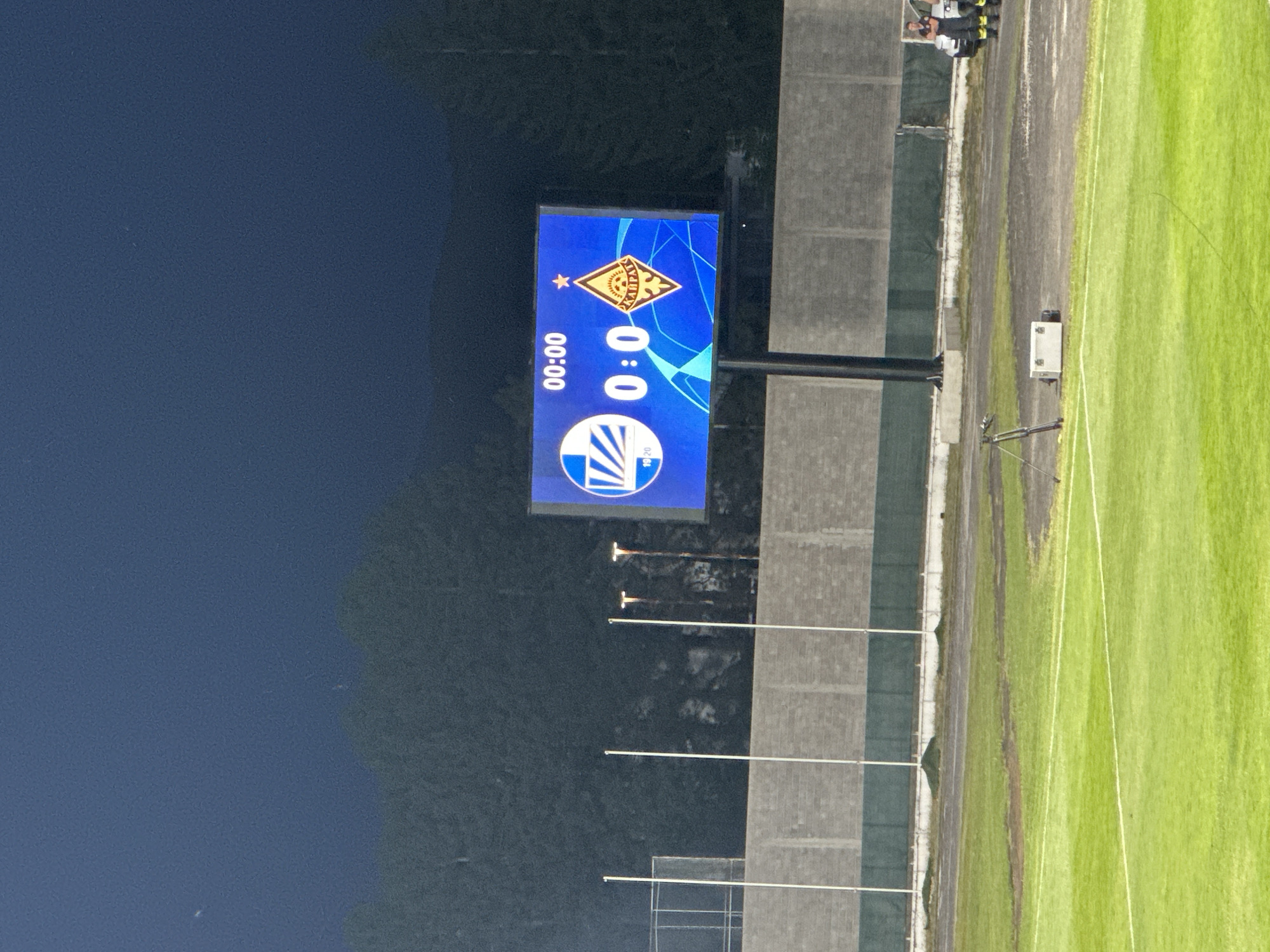The image depicts a nighttime scene at a football stadium, likely in Nikšić, Montenegro, given the context. A large electronic scoreboard displays "00:00" for the time and "0:0" for the score, indicating the start of a match or a game that has not yet begun or has been paused. Two team logos are visible on the scoreboard: one on the left with the number "19 20" below it, and another on the right, a diamond-shaped emblem with a star on top, and the word "KARPAT" prominently displayed within.
The stadium environment includes a well-maintained green football pitch with white lines. In the background, a grey brick wall separates the pitch from a track or pathway. Behind the wall, several tall white poles are erected, some with lighting fixtures attached. Further in the distance, dense trees form a dark backdrop against the night sky.
On the right side of the image, two individuals in what appear to be firefighter uniforms are standing near a white vehicle, possibly providing support or security for the event. In the foreground, a portable camera tripod and a white equipment case are placed on the ground near the edge of the pitch. The overall atmosphere is dark, illuminated by the stadium lights and the bright scoreboard.