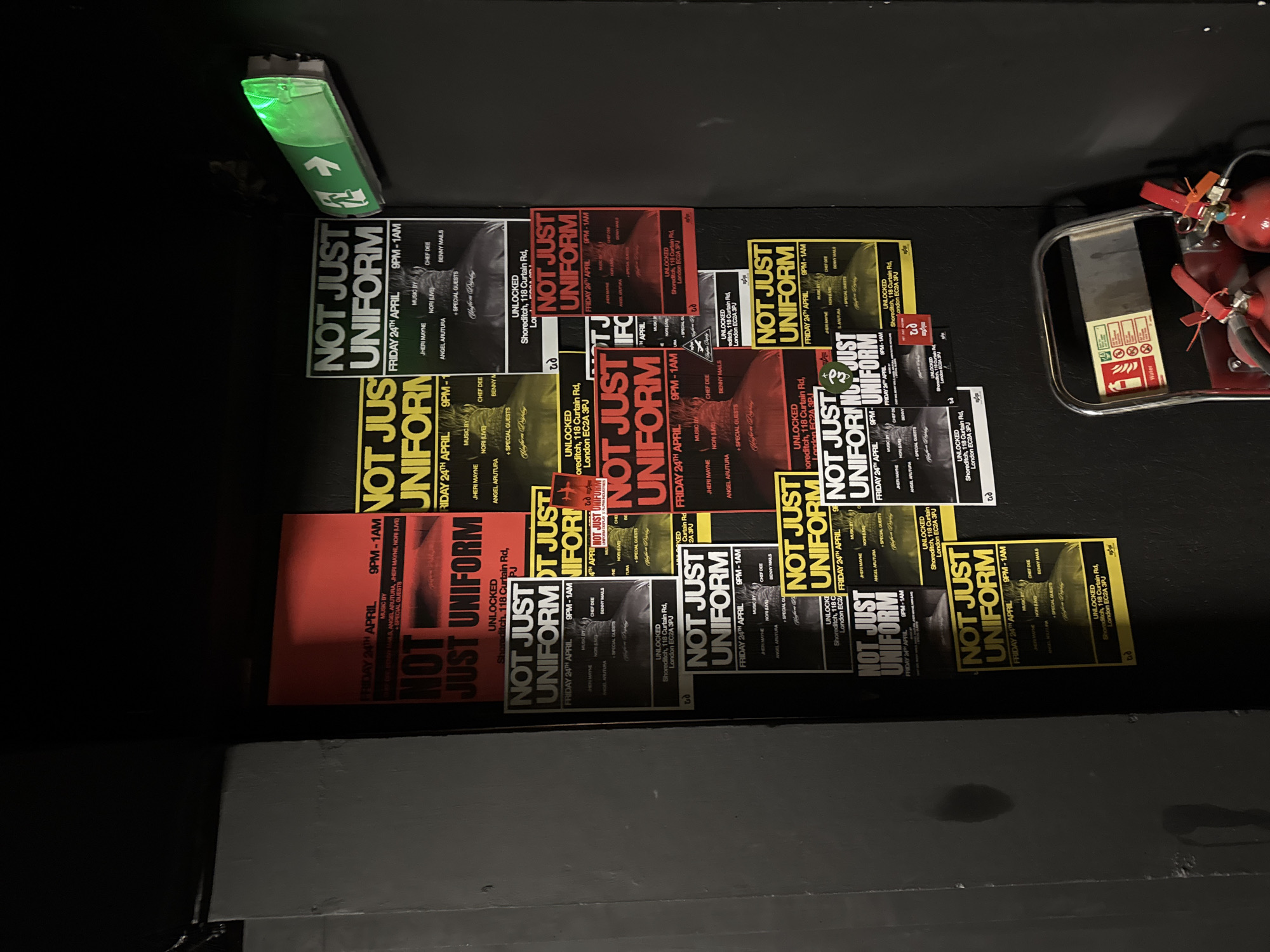 The image displays a collection of numerous event posters, primarily for an event titled "NOT JUST UNIFORM," affixed to a dark wall or floor surface. The posters vary slightly in color scheme, featuring red, yellow, and black designs, but share common information.
The event, "NOT JUST UNIFORM," is scheduled for "FRIDAY 24TH APRIL," with times typically listed as "9PM - 1AM," though one poster indicates "8PM - 1AM." The location is specified as "UNLOCKED Shoreditch, 118 Curtain Rd, London EC2A 3PJ," confirming a London, United Kingdom setting. The posters list several artists and DJs providing music, including "JHERI MAYNE," "CHEF DEE," "NORI (LIVE)," "ANGEL ARUTURA," and "BENNY MAILS," along with "+ SPECIAL GUESTS." Some posters also have a small logo or text that appears to say "Uniform Display" or similar.
Above the posters on the left, a green emergency exit sign is visible, illuminated and pointing upwards with a running person pictogram. To the right of the posters, two red fire extinguishers are mounted on a metal trolley or cart. A small label on the cart shows fire safety pictograms, including one indicating "Water." The overall scene suggests an indoor venue, likely a club or event space, given the dark surroundings, event posters, and safety equipment. The perspective is a slight aerial view, looking down at the arrangement of posters.