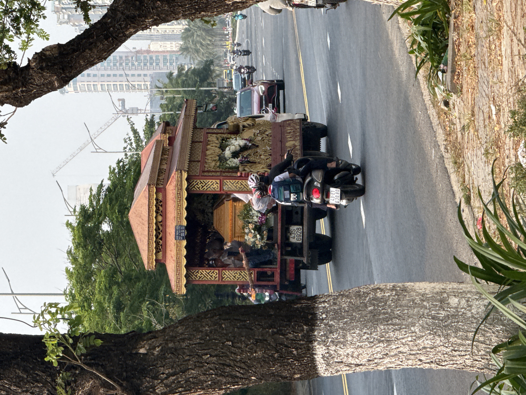 A slow-moving procession travels along a coastal road in Đà Nẵng, Vietnam. The scene is framed by trees on either side of the road. The focal point is an elaborate, ornately decorated structure mounted on a flatbed truck. Inside the structure are several people dressed in casual clothing. The structure is also adorned with flowers.
A motorcycle follows closely behind the truck. In front of the truck, other vehicles and motorcycles are also visible. Tall buildings are visible in the distance. The road has yellow lines dividing traffic lanes.