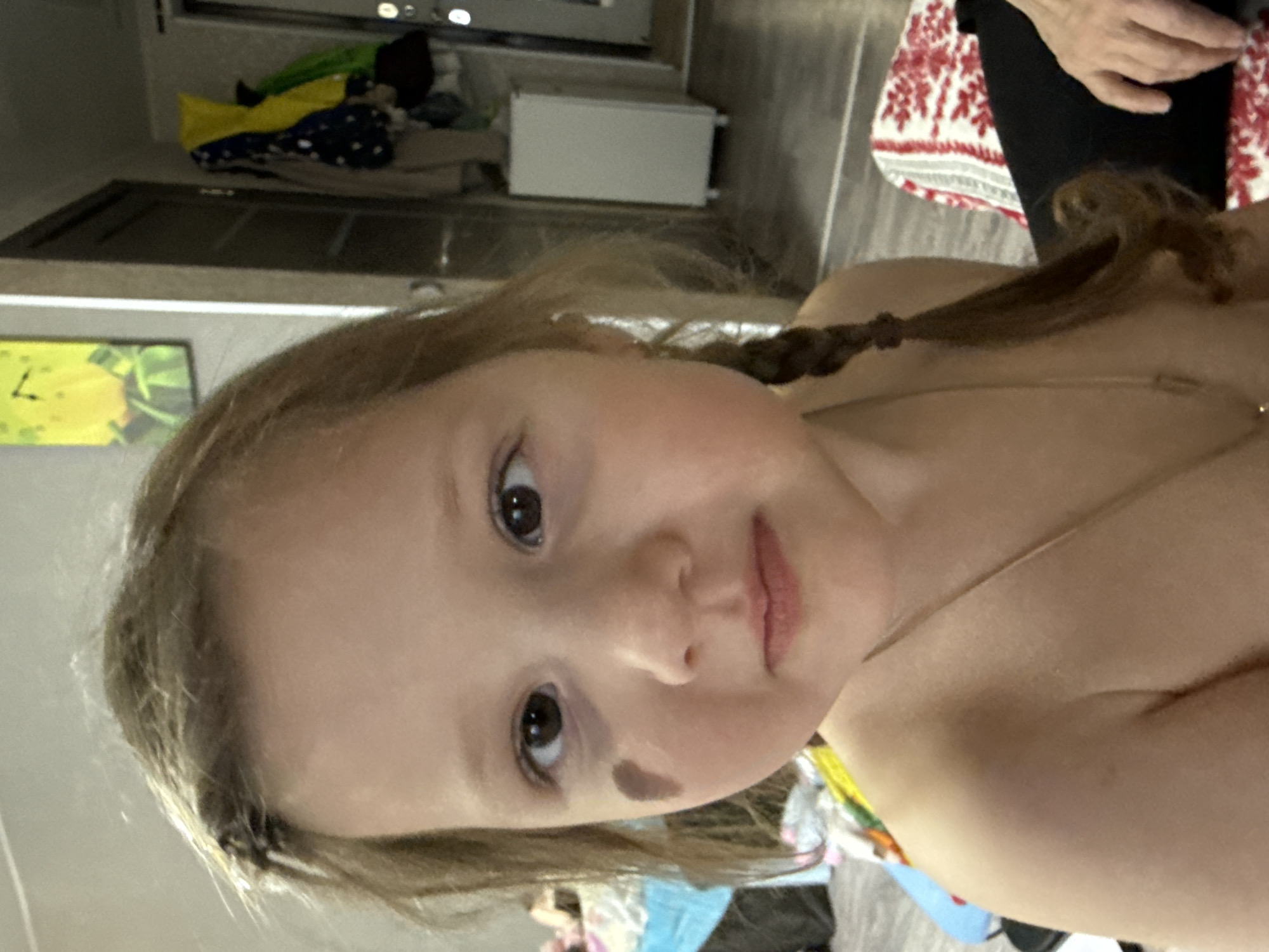 A close-up shot depicts a young girl with light brown hair, styled with a small braid. She has fair skin and brown eyes, and a small mole is visible on her left cheek. She appears to be wearing a thin necklace. The background is cluttered, showing a glimpse of clothes, a possible towel, and an indeterminate room setting. The location in the city of Кипчак, Russia, cannot be determined from the visual information provided.