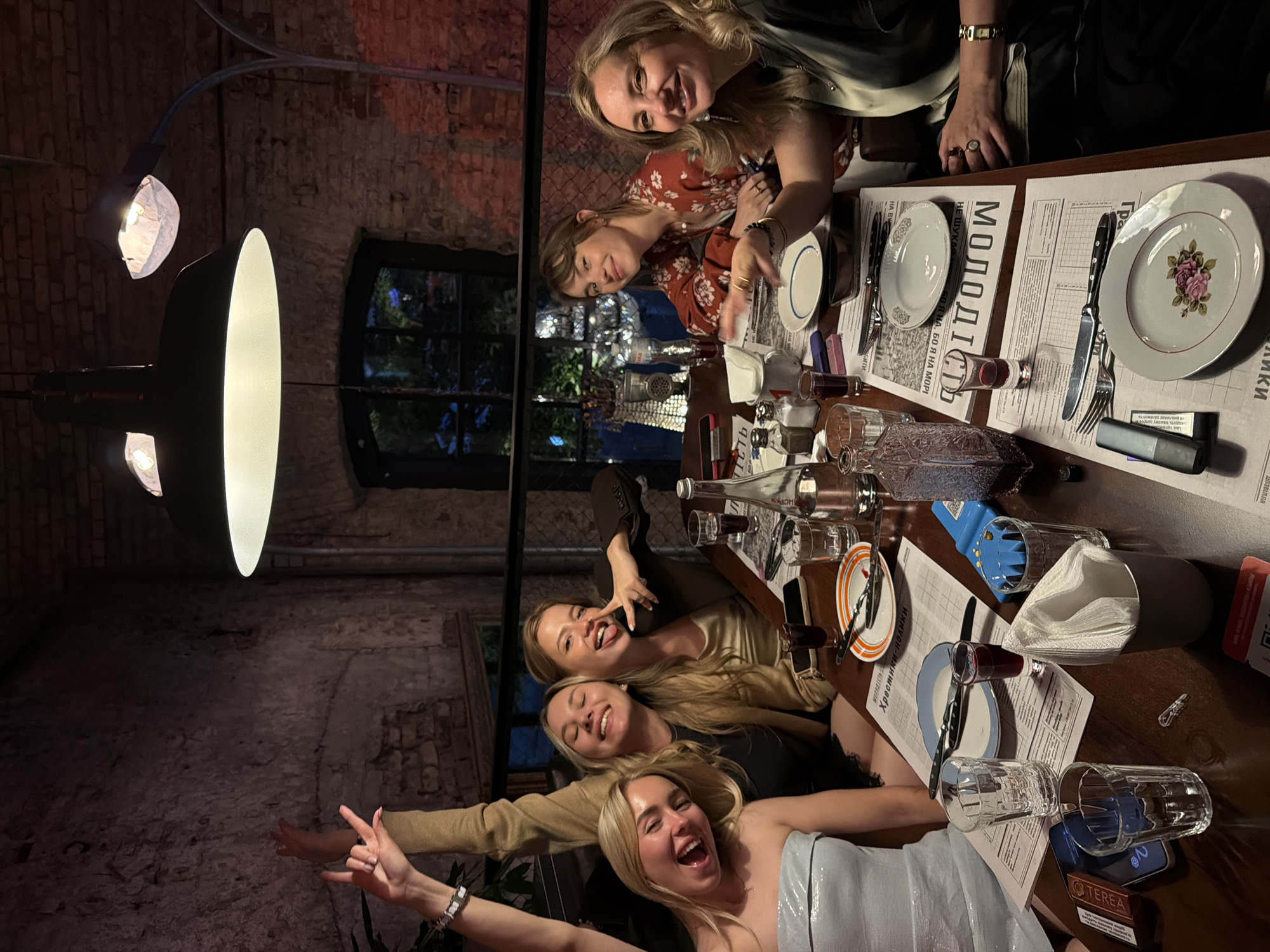 Six women are dining at a table in a dimly lit restaurant with exposed brick walls. The table is set with plates, glasses, silverware, and napkins. Some of the women are smiling and laughing, and one is making a peace sign with her fingers. The woman on the far left has her arm raised in the air with her hand in a rock-on gesture. The woman second from the left is wearing a tan jacket and has her arm raised as well. The woman in the foreground on the right is wearing a black and white striped top and a black skirt. The woman to her left is wearing a red floral top. The woman in the foreground on the left is wearing a sparkly silver strapless dress. A menu or newspaper is laid out on the table. The restaurant appears to be in Kyiv, Ukraine, based on the Cyrillic script visible on the table in multiple locations.