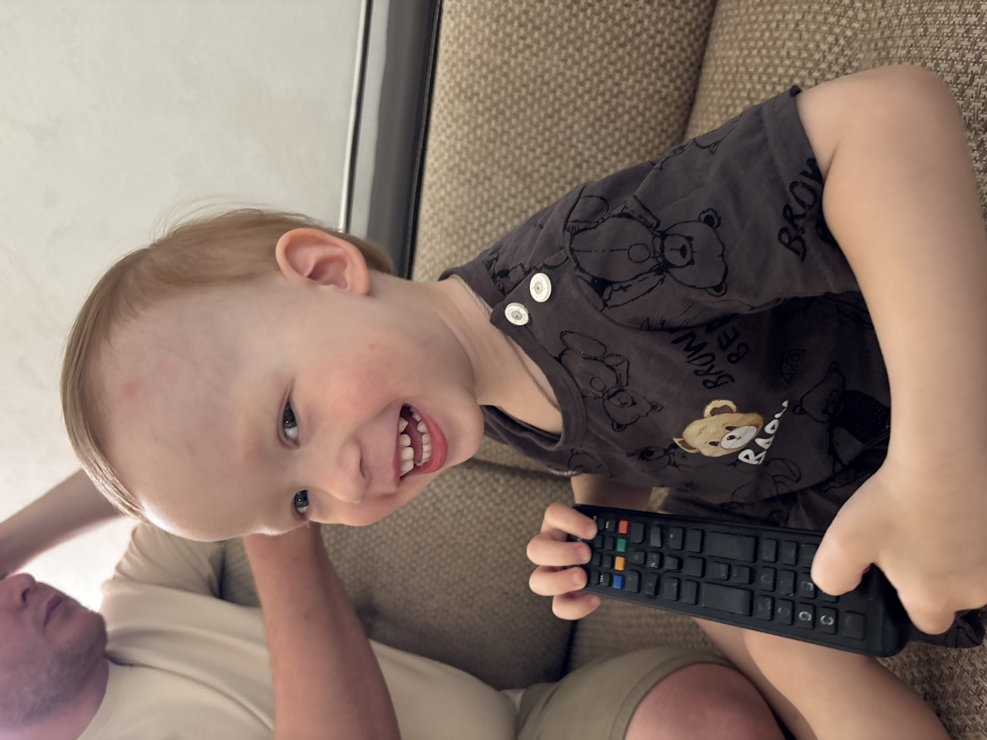 A young child with fair skin and blonde hair is sitting on a light brown couch and holding a black remote control. The child is wearing a dark gray t-shirt with teddy bear graphics and white buttons. Part of an adult is visible in the lower left corner of the image, including their face and arm. The adult is wearing a light-colored shirt. The location is likely indoors. The city of Kochkino, Russia, is not discernable from the image.