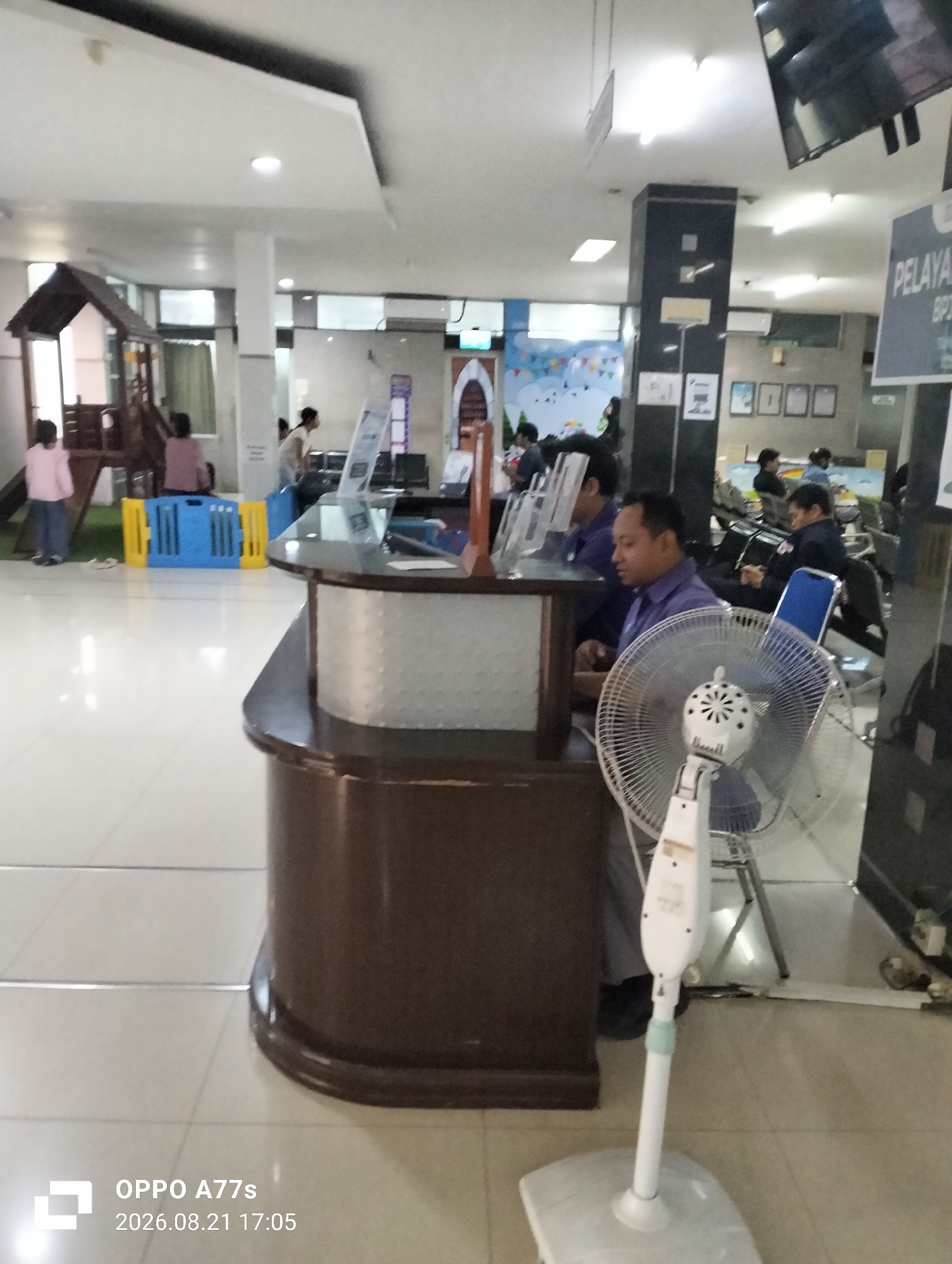 The image depicts the interior of a public service area, possibly a government office or a hospital waiting room, in Semarang, Indonesia. In the foreground, a dark brown counter with a white textured panel is visible. A white oscillating fan is positioned in front of the counter, with its blades turned off. Behind the counter, three men are seated, appearing to be employees, and one man is standing. The man in the center is wearing a blue uniform shirt, as are the other two seated men, suggesting they are service personnel.
To the left of the counter, a wooden children's play structure is situated, with two young girls in casual clothing standing near it. One girl wears a pink long-sleeved shirt and dark pants, while the other is partially obscured and wears a pink shirt and dark pants. A blue and yellow plastic barrier is placed between the play area and the service counter.
In the background, several individuals are seated in chairs, seemingly waiting. A sign with the text "PELAYAN" is visible on the right side of the image, which translates to "Service" in Indonesian. The environment appears to be well-lit with overhead lights. The floor is tiled with a light-colored, reflective material. The overall atmosphere suggests a busy, functional public space. The timestamp "2026.08.21 17:05" is superimposed on the bottom left corner of the image, indicating the date and time of capture, and "OPPO A77s" likely indicates the device used.