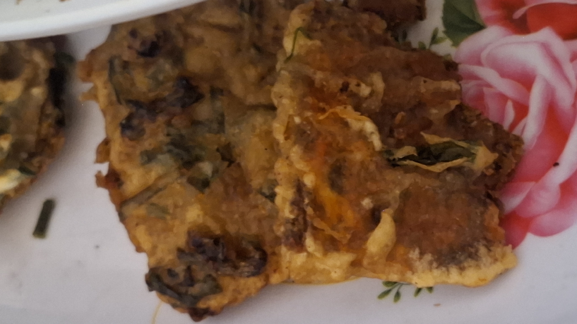 The image shows a close-up of two pieces of fried food on a plate with a floral pattern. The food appears to be some kind of fritter or pancake, with visible green herbs or vegetables mixed into the batter. The texture is slightly crispy on the edges and softer in the center. The plate has a pink and white floral design. The image is taken from a slightly elevated angle, focusing on the food. There are no people or clear environmental context within the frame to indicate a specific location in Cibinong Hilir, Indonesia.