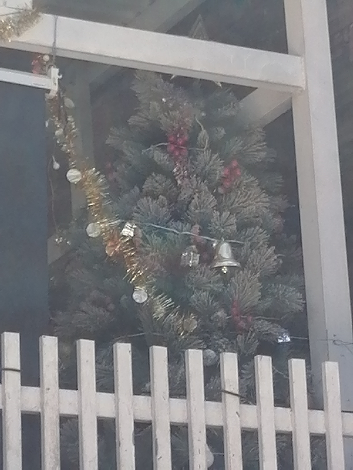 The image shows a Christmas tree decorated with red baubles and tinsel, as well as a silver bell. A white picket fence is in the foreground. The tree appears to be located outdoors, possibly in a yard or public space, given the fence. The scene is viewed through what appears to be a window or some sort of enclosure, indicated by the white frame at the top of the image. The lighting suggests it is daytime. The image's origin in Da Lat, Vietnam, is noted.