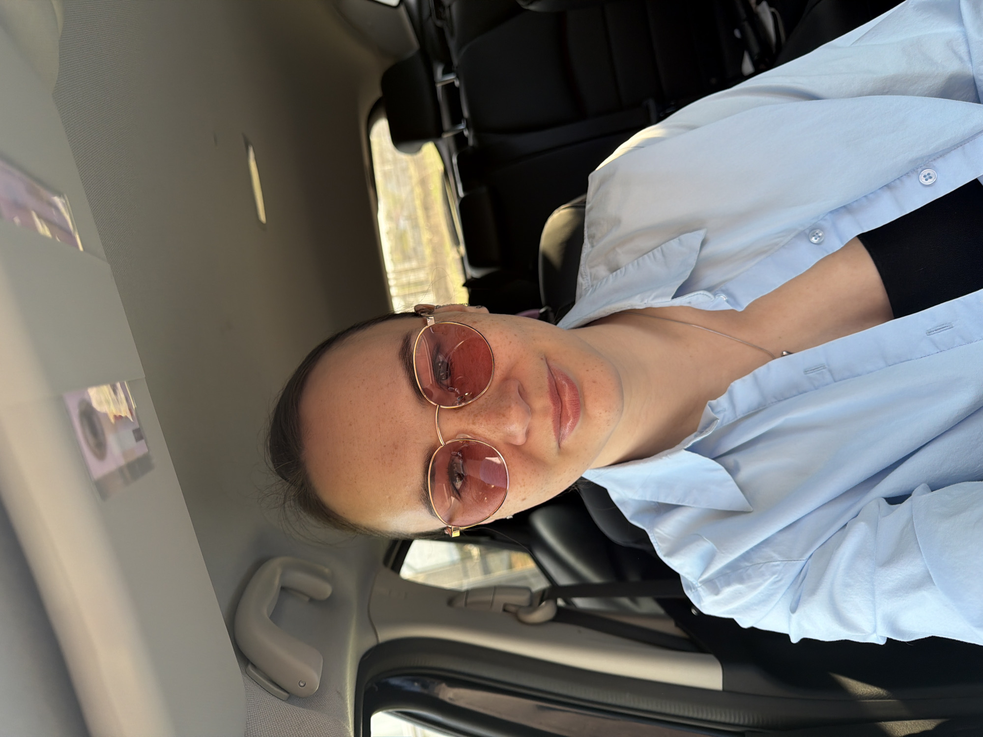 A woman is in a car, wearing rose-tinted sunglasses and a light blue button-down shirt over a black top. The interior of the car is visible. The location is implied to be Chaikovsky, Russia, though this is not visible in the image.