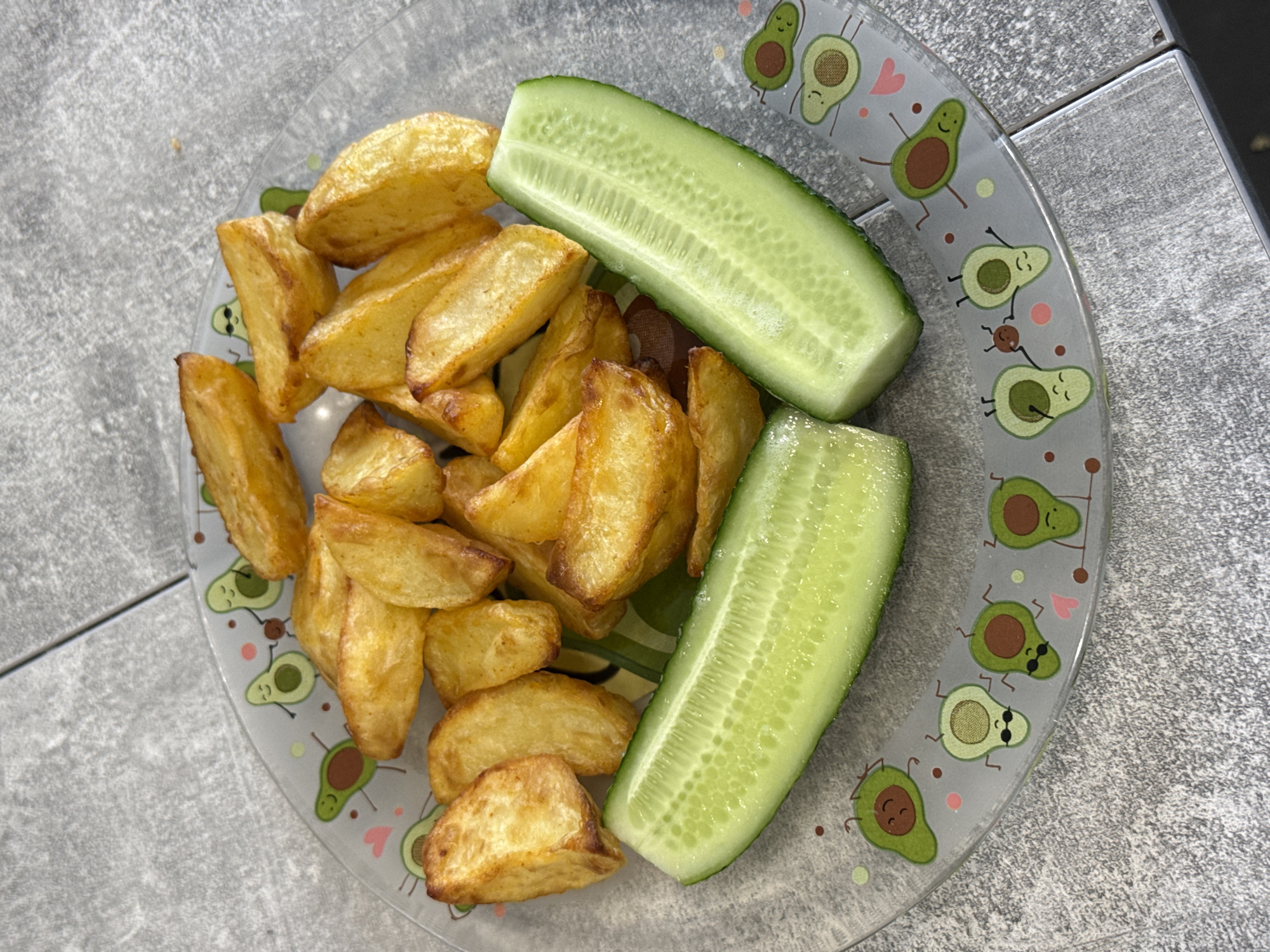 A top-down shot shows a clear glass plate filled with golden potato wedges and two halves of a cucumber on a gray, tiled surface. The plate is decorated with cartoon avocados. The scene is indoors, likely a kitchen or dining area, with the gray tiles suggesting a modern aesthetic. There are no people or animals visible. The lighting is natural, indicating daylight. The image does not provide any information about the city of Lyubertsy, Russia.