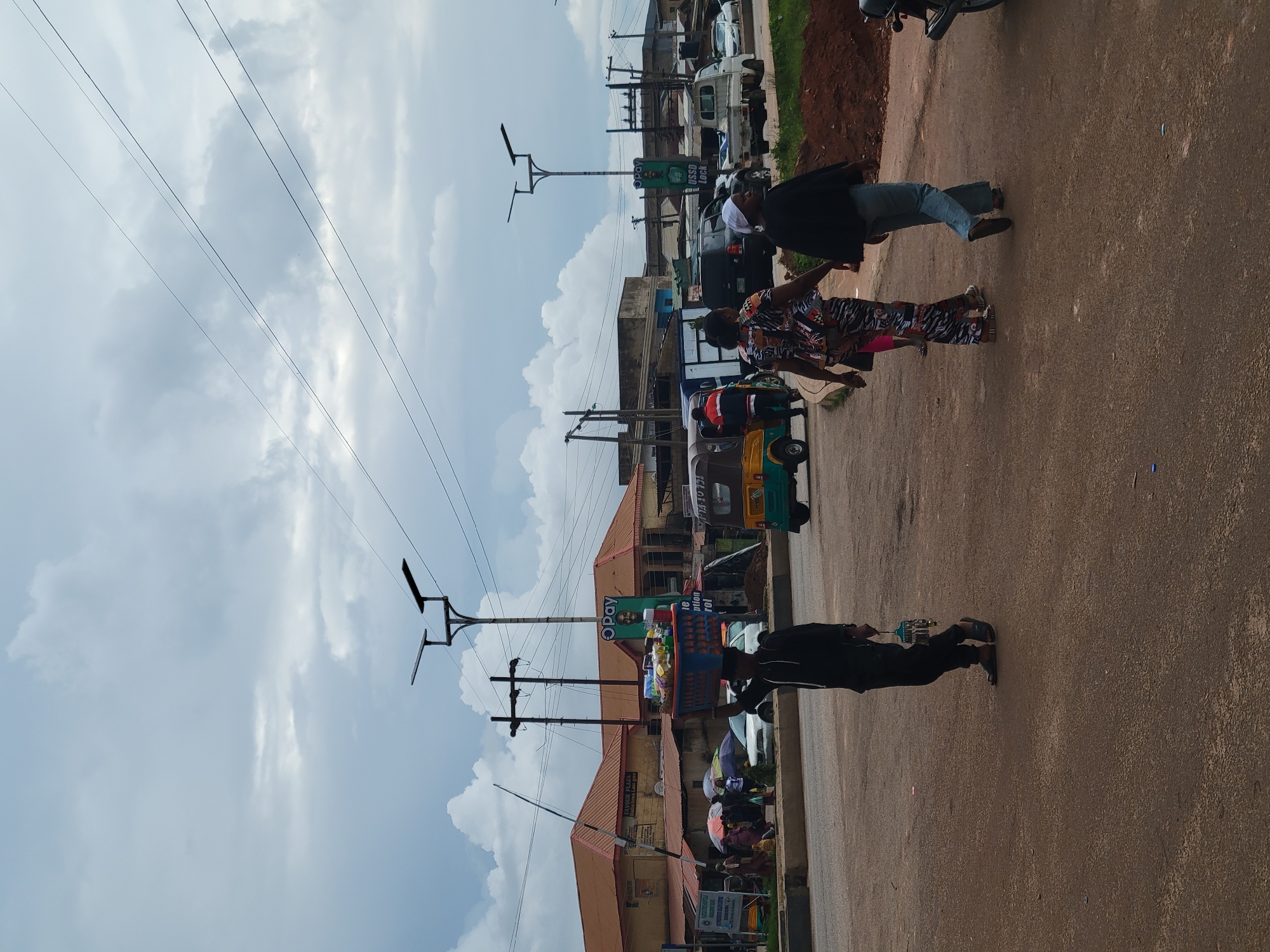 The image depicts a street scene in Gura Top, Nigeria, on a cloudy day. In the foreground, a young man walks from left to right, carrying a large blue basket filled with various items, including food products and other merchandise, balanced on his head. He is dressed in a black hoodie and dark pants, with sandals on his feet. To his right, two women walk in the same direction. The woman closer to the foreground wears a black top and light blue jeans. The woman in front of her is dressed in a colorful, patterned outfit. Behind them, a yellow and green motorized tricycle, commonly known as an "Okada" or "Keke Napep," is parked.
In the background, a street lined with buildings with reddish-brown roofs is visible. Some of these buildings appear to be shops or businesses, with signage including "FAVOUR PLAZA" and an "OPay" advertisement. Several utility poles with power lines and solar street lights are present, indicating infrastructure within the town. Other vehicles, including cars and a small truck, are also visible on the road, suggesting moderate traffic. Umbrellas are deployed by some pedestrians near the shops, possibly to shield from the sun or anticipated rain. The ground is paved with asphalt, and there is a pile of red earth on the right side of the image, suggesting ongoing construction or landscaping. The sky is filled with large, gray clouds, creating an overcast atmosphere.