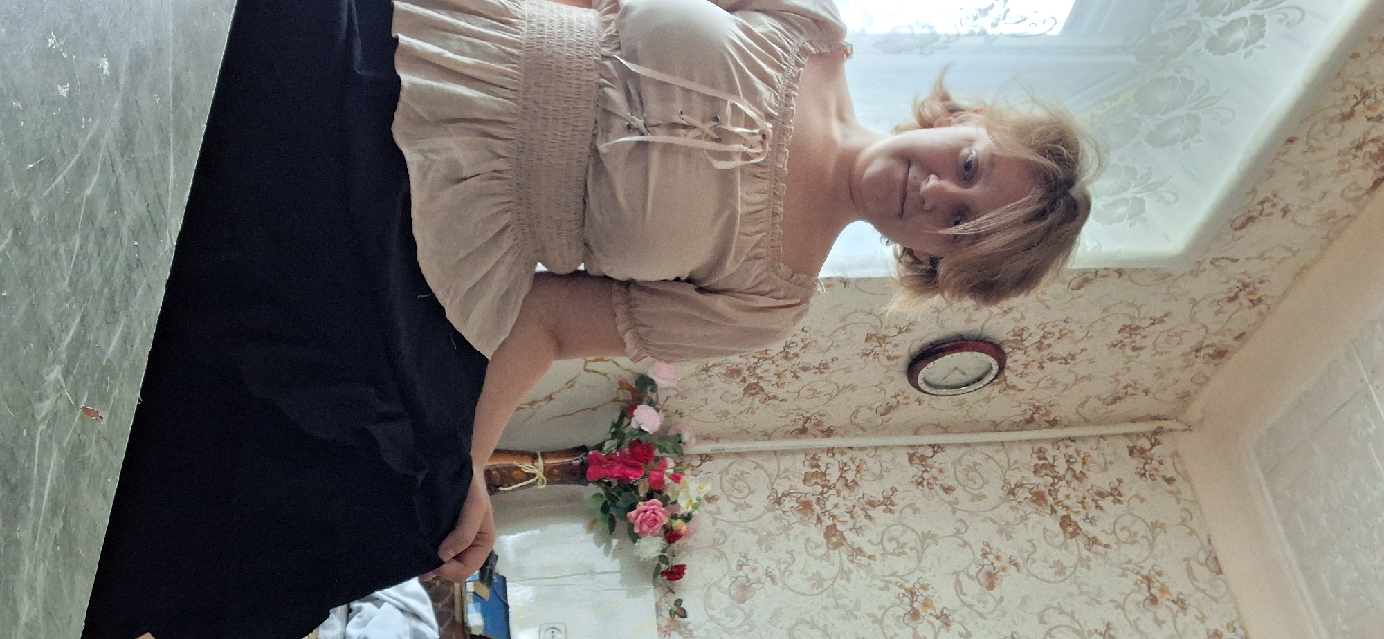 A woman with shoulder-length, light brown hair sits in an indoor setting. She is wearing a light beige, off-the-shoulder blouse with puffed sleeves and a cinched, smocked waist, paired with a long, dark navy or black skirt. Her left arm is resting on her lap, and her right arm is bent, with her hand resting on her right knee.
The background consists of patterned wallpaper with floral motifs in cream, brown, and muted orange tones. To the left of the woman, a window with sheer white curtains is visible. On the wall behind her, a round clock with a dark wooden frame is mounted. To her right, a vase containing a bouquet of artificial flowers in pink, red, and white is placed on a surface. The foreground shows a flat, reflective surface, possibly a table or countertop, with a marble-like pattern. The overall lighting suggests an indoor environment, possibly a room in a residential building, which is consistent with the typical interiors found in Petropavl, Kazakhstan.