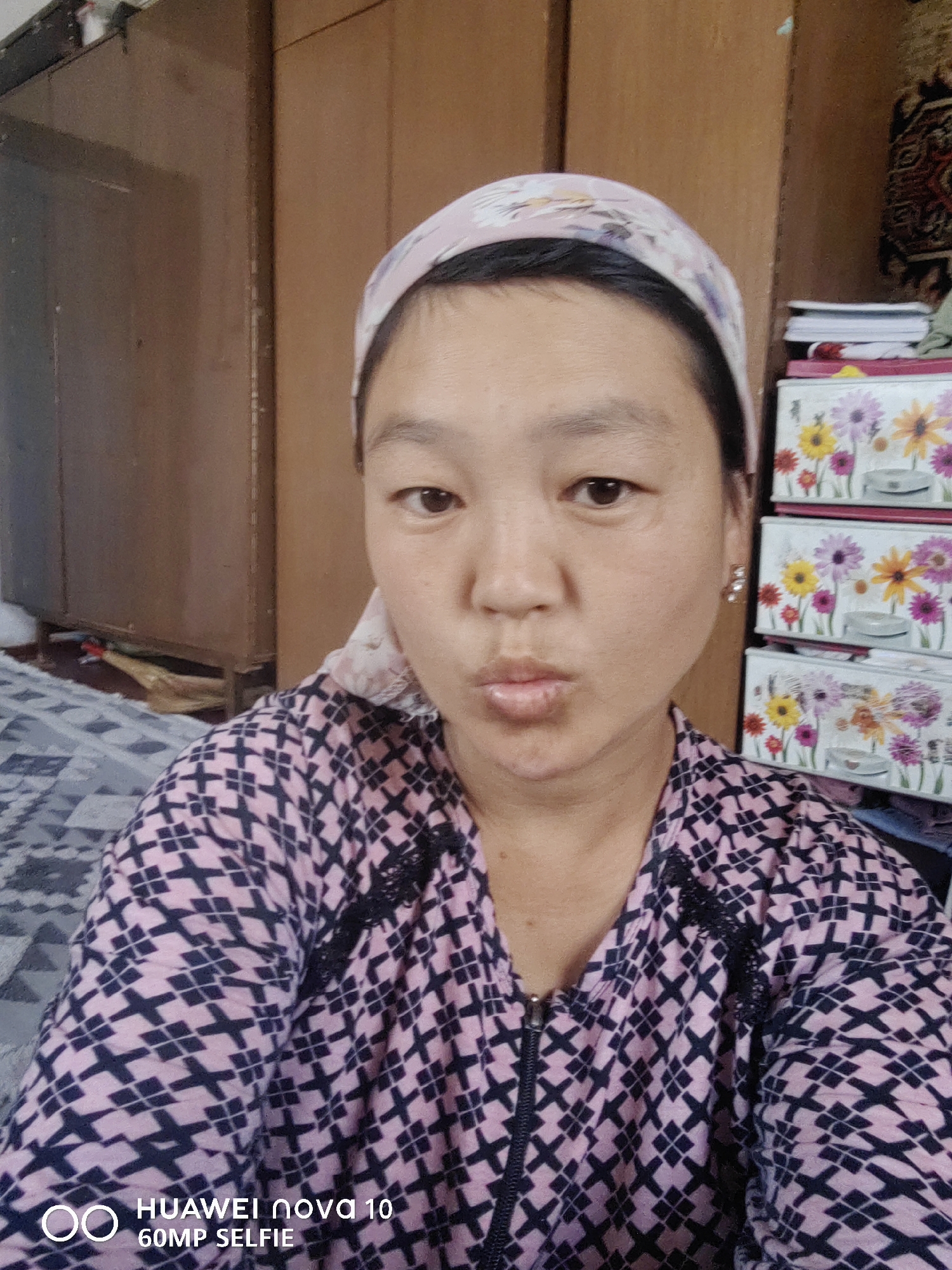 The image shows a medium shot of a woman in Jetim-Dobo, Kyrgyzstan. She is wearing a pink and navy patterned top and a light pink headscarf. She has her lips pursed and is looking towards the camera. In the background, there are wooden cabinets and a stack of colorful boxes. The lighting is natural, suggesting the photo was taken indoors during the day. The overall atmosphere appears casual and domestic.