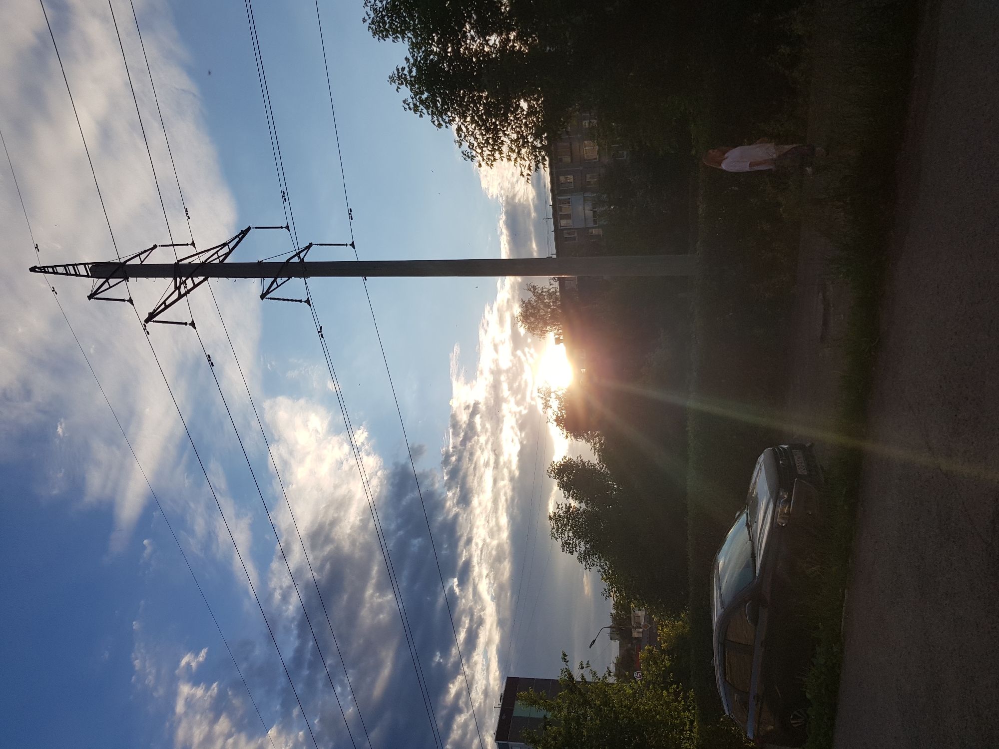 In a silhouetted scene at dusk in an urban environment, a dark-colored station wagon is parked on the side of a paved road, with its front facing left. Green foliage borders the vehicle. Further to the right, a person in a white top and patterned pants walks away from the camera. Power lines crisscross the sky, supported by a tall, industrial pylon. In the background, trees and apartment buildings are visible against a sky with dramatic clouds and a sunburst effect from the setting sun. The overall lighting suggests early evening.