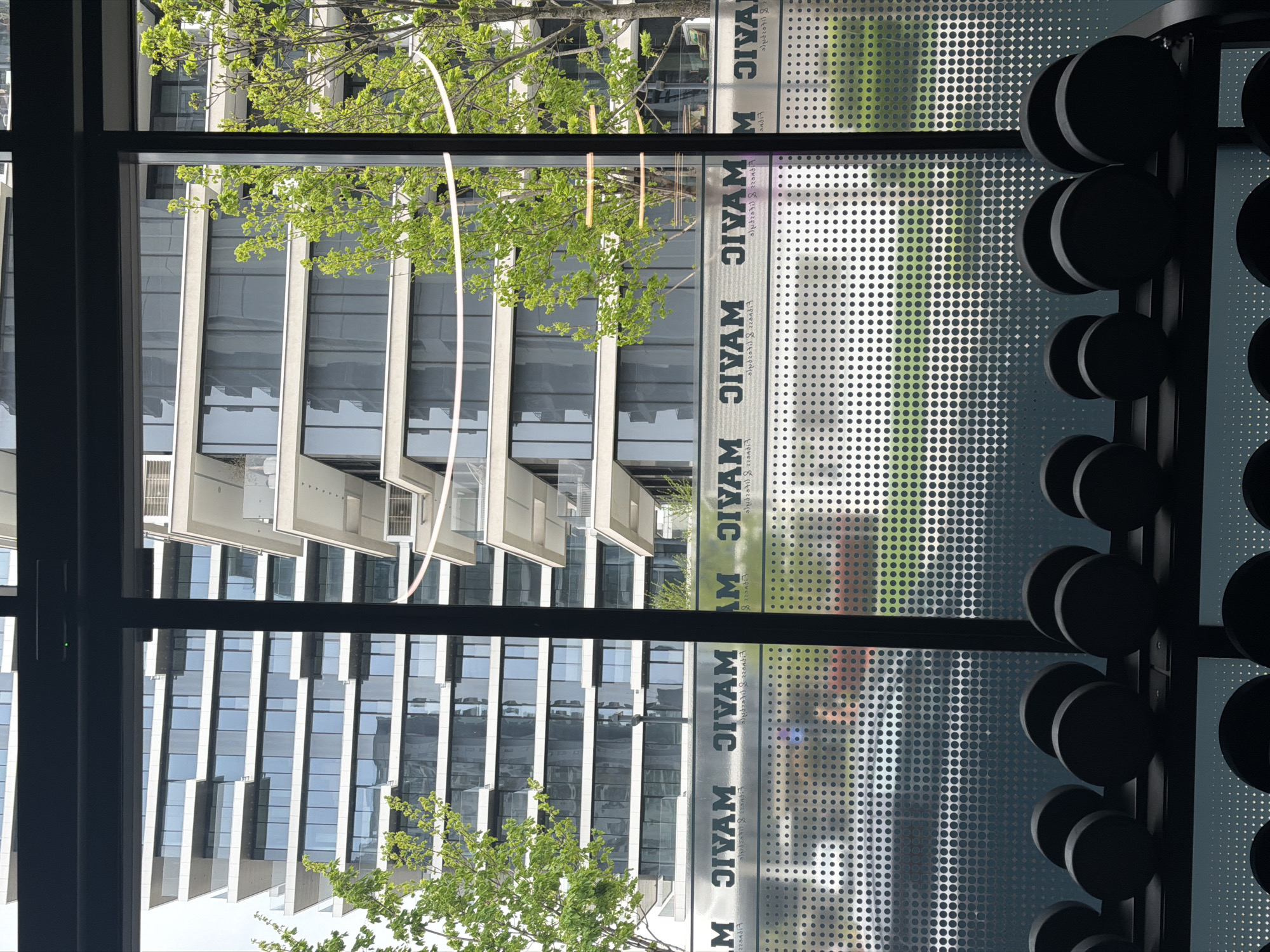 The image shows a view through a window with a black frame. Outside, there is a modern building with a series of windows and protruding balconies. A tree with green leaves is partially visible in the foreground, obscuring some of the building. The window has a decorative pattern of small circles and some text that appears to be mirrored. To the right of the window, there is a rack of dumbbells.
I cannot give you a location in the city of Belgrade, Serbia, because it is not specified in the image.