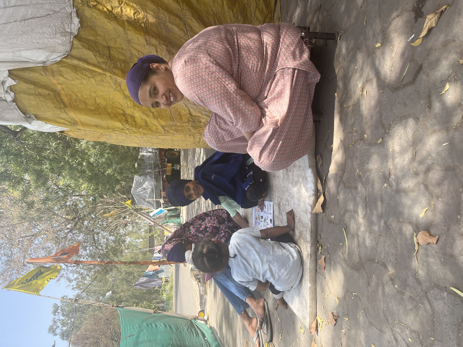 Four people are sitting on a paved area outside. They are sitting on a white, rectangular sheet. Three of them are wearing turbans, and one is wearing a pink floral dress. One of the women is wearing a blue turban. The other three are wearing blue turbans. The woman wearing the pink floral dress is sitting on a small, metal stool. One of the women is looking at the camera and smiling. The people are in the city of Sahibzada Ajit Singh Nagar, India. There is a large yellow tarp behind the people. To the left of the people is a green tarp and an older woman wearing a dark, floral print headscarf is partially visible. She is wearing a light blue shirt and is holding what appears to be a book. A young boy is sitting in front of her and appears to be playing with a book. There are several trees and a building in the background.