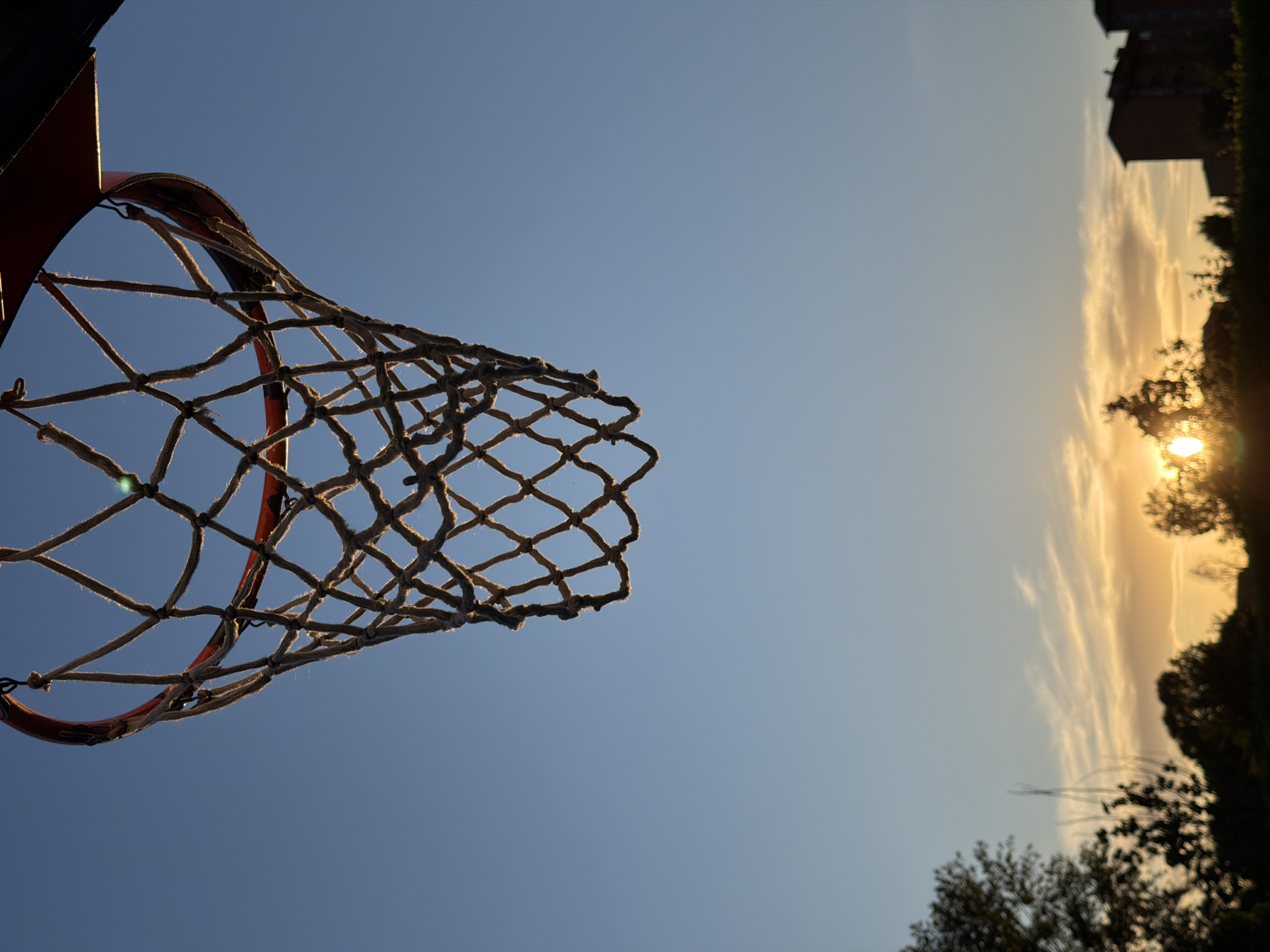A low-angle shot captures a basketball hoop with a rope net against a blue and yellow sky at dusk. The hoop is positioned on the left side of the frame, with the setting sun visible on the right, partially obscured by trees. A building is also faintly visible in the upper right. Although I can not confirm the location, the prompt indicates this photo was taken in the city of Milan, Italy.