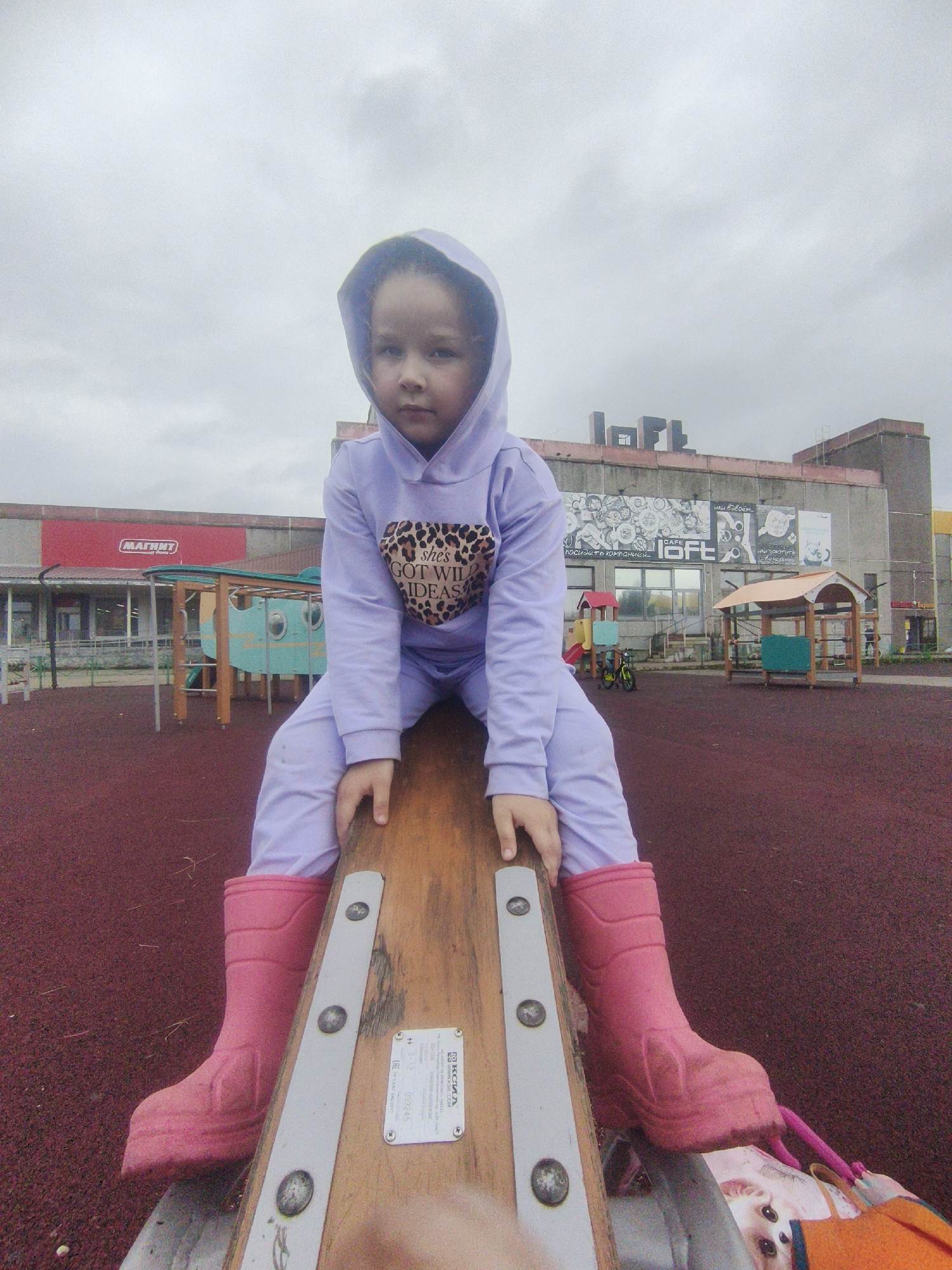 A young girl with dark hair, wearing a purple hooded tracksuit with leopard print text that says "she's got wild ideas" and pink rubber boots, sits on a wooden seesaw in a playground. She is facing the camera. The playground surface is red rubber mulch. In the background, there is a building with a red sign that says "МАГНИТ," a children's play structure, and a building with graffiti and signs that read "CAFE LOFT." The sky is overcast. The location is the playground in Markovsky, Russia.
