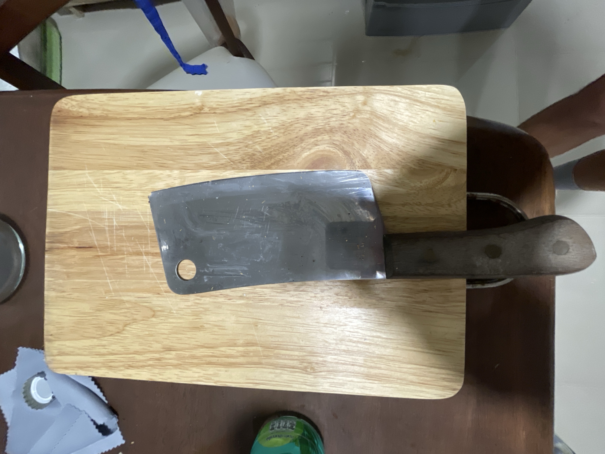 The image shows a wooden cutting board with a cleaver knife on top, positioned on a dark brown wooden table. A green bottle with some writing is visible on the left side of the table, partially obscured by the cutting board. A grey cloth with a bottle cap is also present on the left. In the background, a white wall is visible with a blue ribbon hanging down. The scene appears to be an indoor setting, likely a kitchen, in Sakhu, Thailand. No people are clearly visible in the image, though a leg and foot in a grey sock are visible at the bottom. The lighting suggests it is daytime.