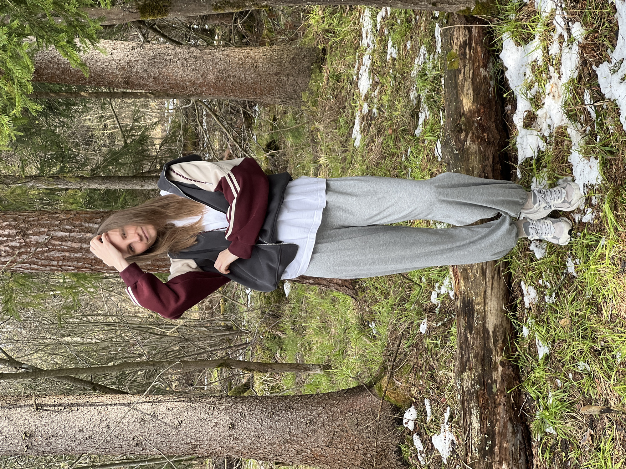 A young woman is standing in a forest. She is wearing grey sweatpants, a white t-shirt, a burgundy and cream-colored jacket, and grey sneakers. She has long brown hair. Her left hand is touching her forehead. She is standing next to a fallen log that has some snow on it. There are trees in the background. The location is Раздолье, Russia.