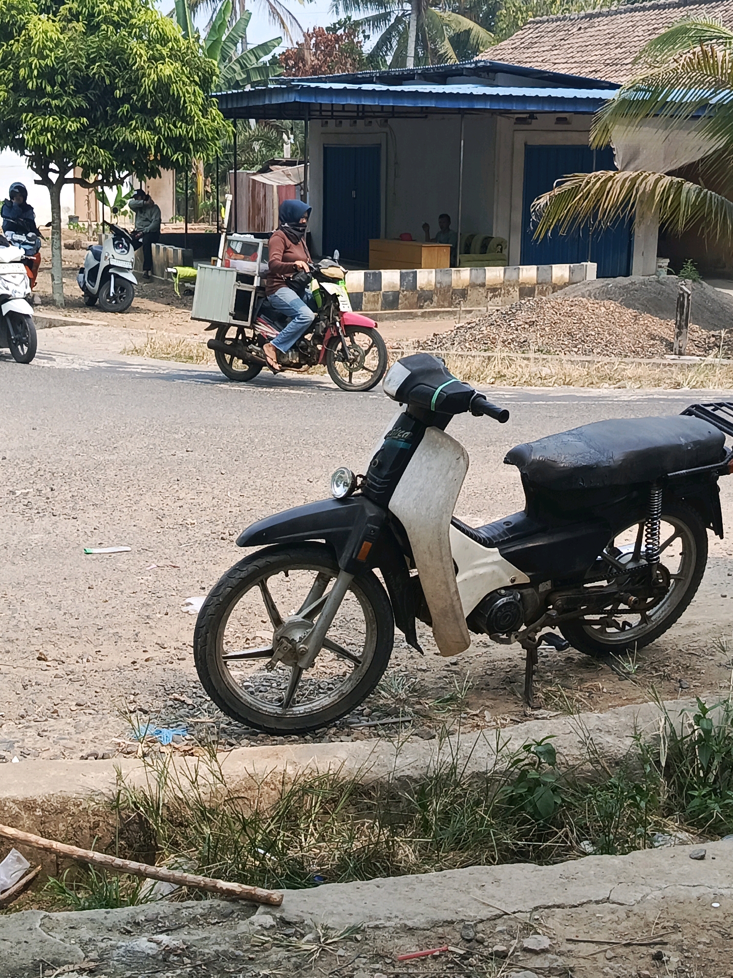 This is a medium shot, eye-level, outdoor photograph taken on a bright, sunny day in Wonosari, Indonesia. The scene depicts a roadside with several motorcycles and a small building. In the foreground, a black and white motorcycle is parked on a dirt and gravel shoulder, facing left. The motorcycle has a black seat and handlebars, a white front fairing, and a black rear fender. To the left of this motorcycle, a portion of another white motorcycle is visible.
Further back, on the paved road, a person wearing a brown jacket, blue jeans, and a dark blue headscarf rides a pink motorcycle. The motorcycle has a white box mounted on the rear. The rider is looking forward. To the left of the pink motorcycle, a white scooter is parked. A person in a gray jacket and a dark blue headscarf is seated on the white scooter, facing forward. Behind them, a person in a blue jacket is standing next to a tree.
The building in the background has a blue corrugated metal roof overhang. It appears to be a shop or stall with a light-colored facade and blue doors. Inside the building, two people are seated on a light green sofa. A pile of gravel and construction materials is visible in the middle ground. The environment appears to be somewhat rural, with a dirt road, sparse vegetation, and trees. A concrete curb runs along the edge of the road in the foreground.