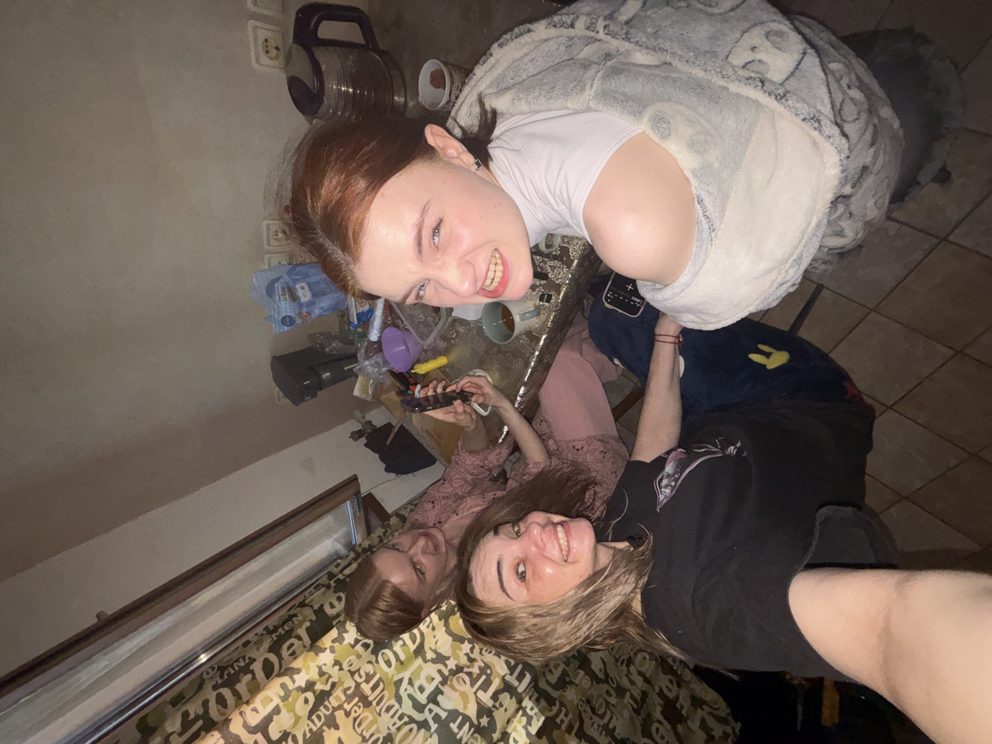 This candid, overhead shot captures three young women relaxing together in what appears to be an indoor residential setting, possibly a bedroom or living room. The artificial lighting suggests it is evening or night.
The woman at the top of the frame has reddish-brown hair and wears a white top under a grey, fluffy blanket or hoodie. She is looking directly at the camera with a wide, cheerful smile, revealing her teeth. To her right, a black and silver vacuum cleaner is visible, along with European-style electrical outlets on a light-colored wall.
In the middle-left, another woman with light brown hair is partially visible, wearing a pink top with teddy bear patterns. She holds a smartphone in her hands, seemingly engaged with it. She offers a subtle smile towards the camera.
At the bottom-right, the third woman, with light brown hair, is also looking at the camera with a broad, happy smile. She is wearing a black t-shirt. Her right arm is visible, and she appears to be lying down on a patterned blanket or bedspread.
Various objects are scattered around a glass-top table, including several cups (one containing a dark beverage like tea or coffee) and a purple funnel. On the phone screen near the top woman, the word "Concert" is partially visible. The patterned blanket features text that includes words like "Order," "Adults," "Men," "LAND," "LANDSCAPE," "UNIVERSE," "STUDIOS," "ENTERTAINMENT," and "WORLD." The floor, visible in some areas, is tiled.
The overall mood is casual and friendly, with all individuals displaying positive emotions through their smiles.