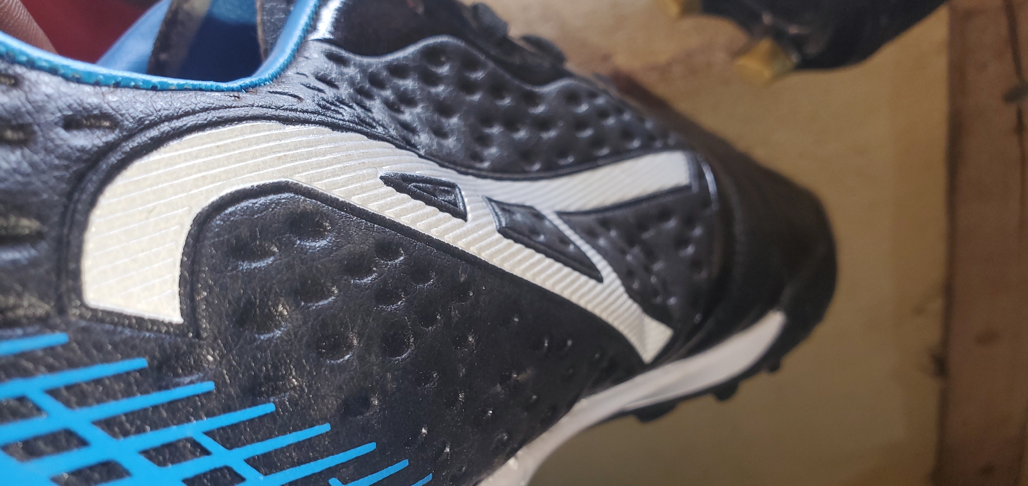 The image shows a close-up view of a black and white athletic shoe with blue accents. The shoe features a textured black upper with a white, stylized "V" or swoosh-like design. The sole of the shoe is not visible, but there are some blue lines on the side, possibly part of the branding or design. The shoe appears to be in good condition. The background is blurry and appears to be a light brown, possibly a floor or surface. The lighting is overhead and somewhat uneven. The image is taken from a high angle looking down at the shoe. There is no indication of the specific location in Bukuru, Nigeria.