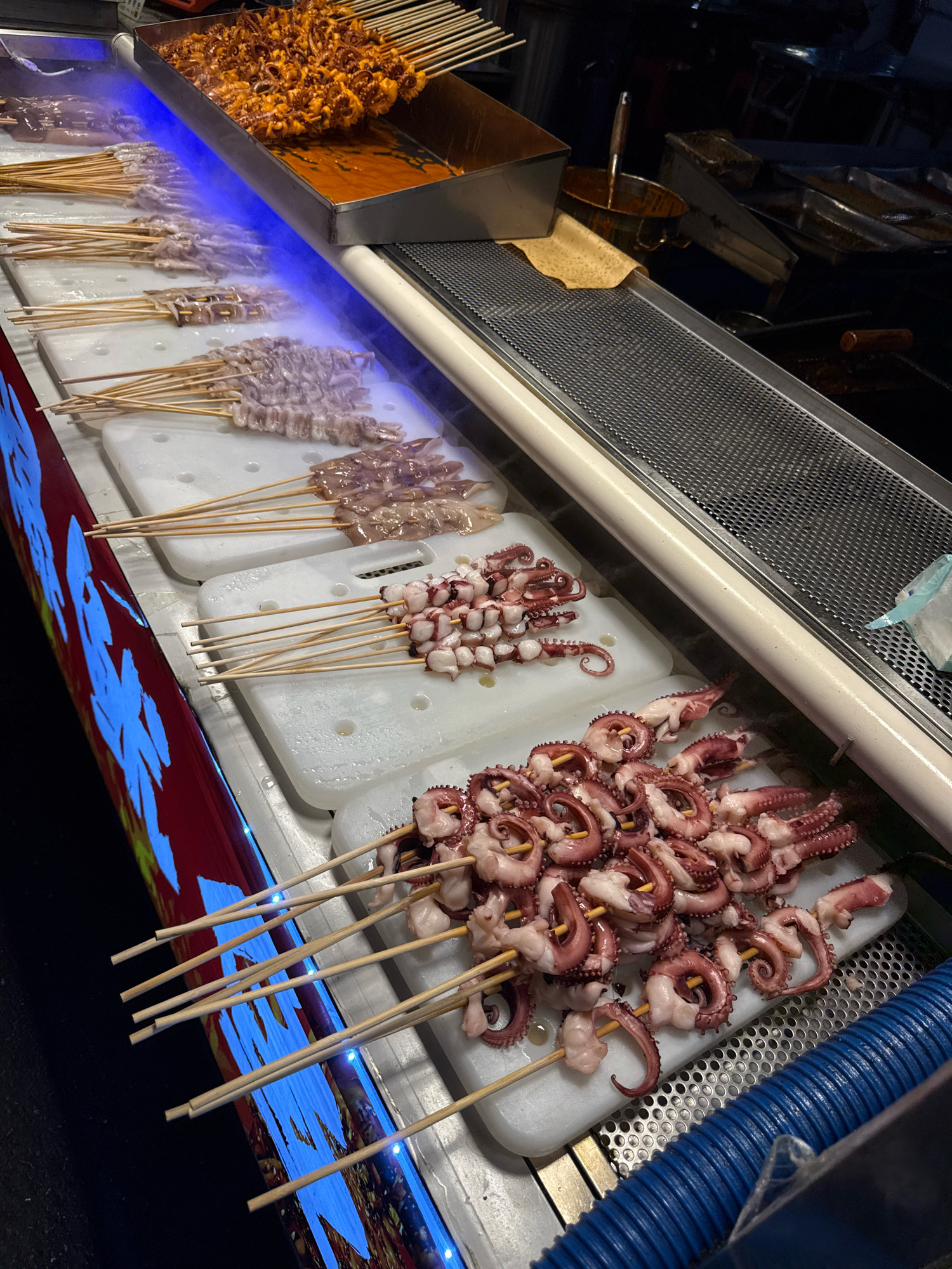 This is a close-up, eye-level shot of a street food stall in Yiwu, China, likely at night given the artificial lighting. The stall features several rows of seafood on skewers displayed on white cutting boards. The most prominent items are rows of small octopuses, their tentacles curled, and what appear to be squid or calamari. In the background, a tray holds a large portion of cooked, reddish-brown seafood, possibly also squid or octopus, covered in a sauce. Blue LED lights illuminate the left side of the display, reflecting off the seafood and the stall's exterior. A metallic grate is visible along the top right, suggesting a cooking or display area. The overall impression is one of a bustling food market, offering a variety of skewered seafood options.