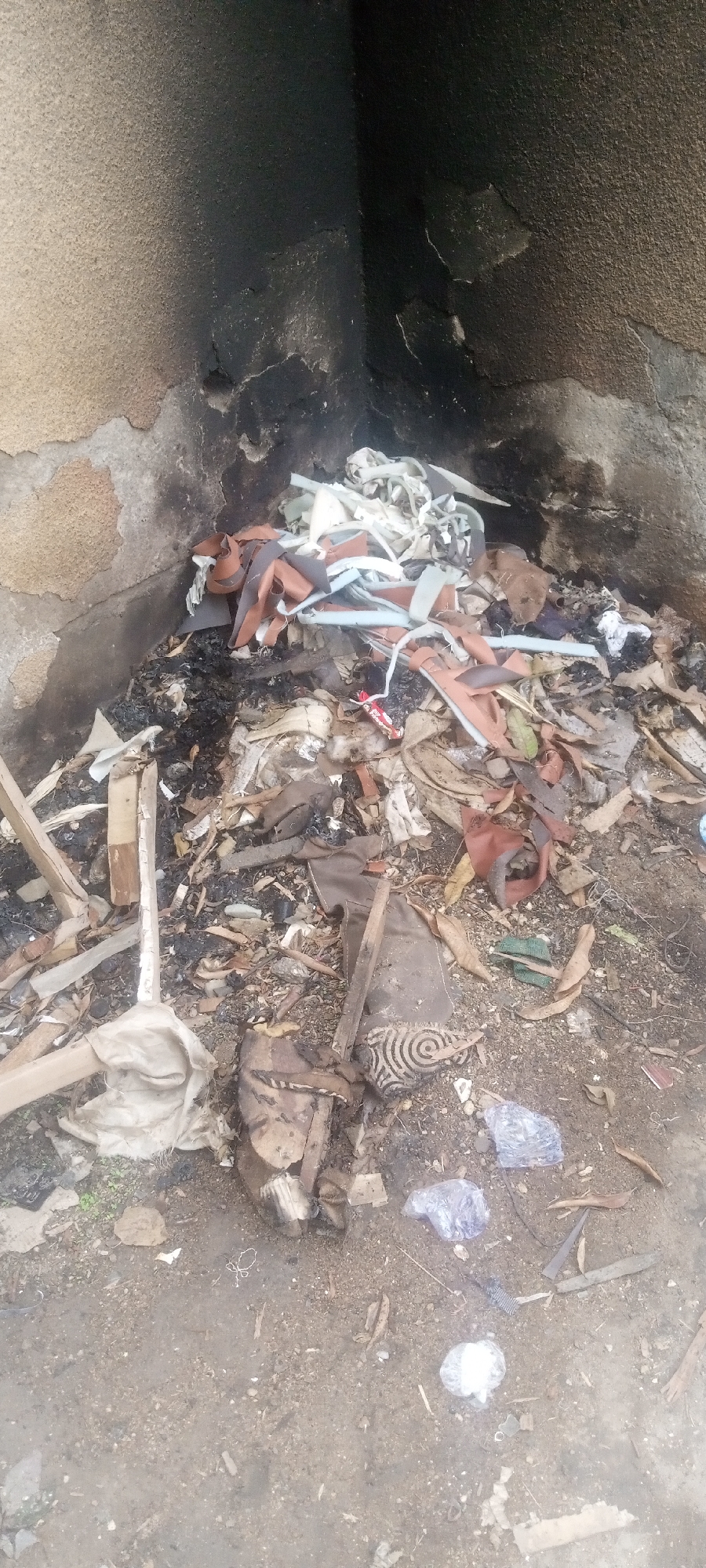 The image depicts a pile of burned and unburned debris located in what appears to be a recessed area next to a building in Sabon Tasha, Nigeria. The wall behind the debris shows signs of fire damage and soot accumulation. The debris includes scraps of wood, fabric, paper, plastic, and what appears to be pieces of furniture or upholstery. The ground is a mix of dirt and small stones. There are no visible people in the frame.