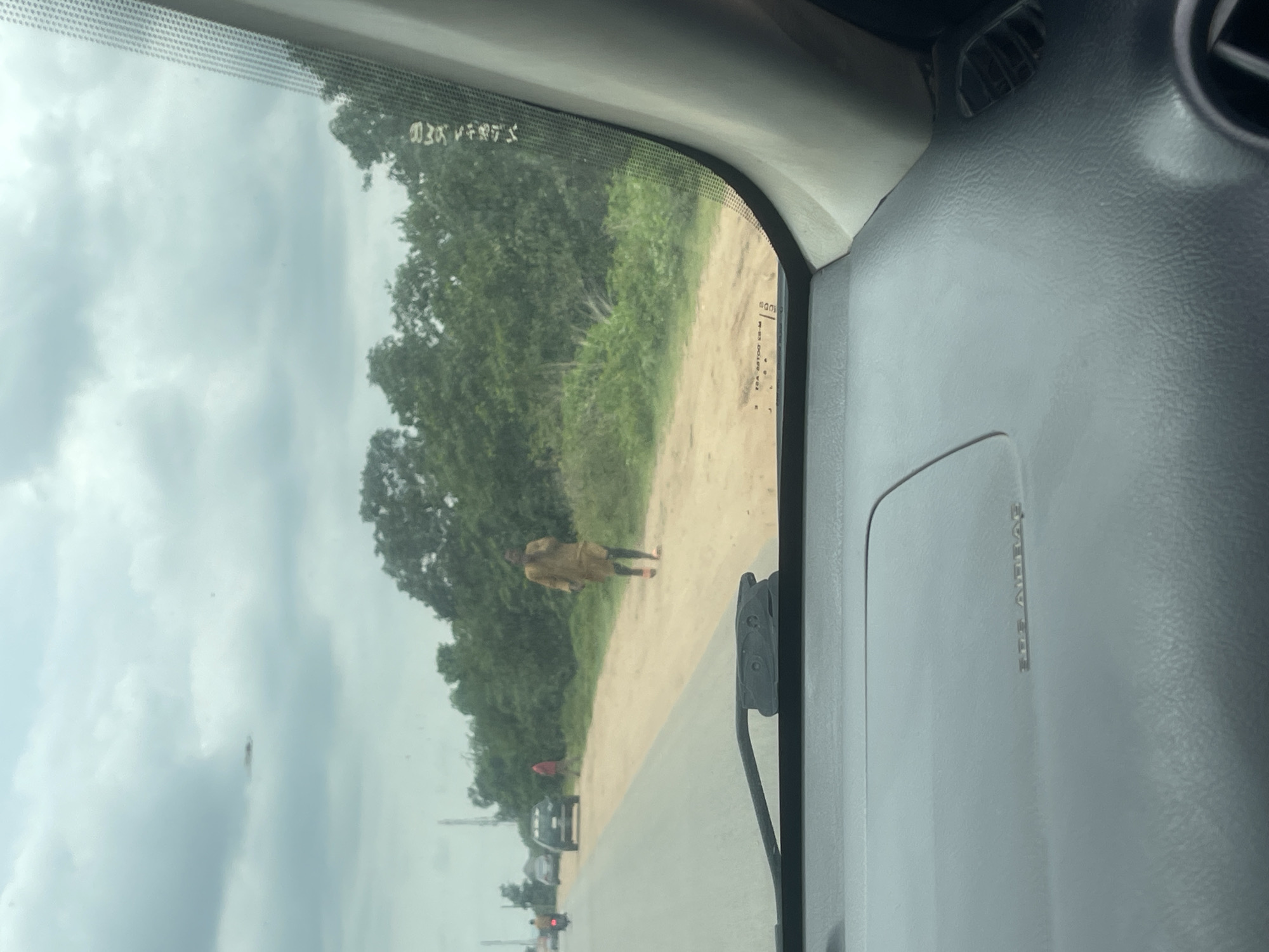 The image, taken from the interior of a vehicle, shows a road scene in Galadimawa, Nigeria. The road is paved and has two lanes visible. On the right side of the road, there is a dirt shoulder with sparse vegetation and trees. A man wearing a tan-colored traditional outfit is walking on the dirt shoulder. In the distance, other vehicles, including a motorcycle and a car, are visible on the road. The sky is overcast with clouds. The foreground of the image shows the dashboard of the vehicle, including a windshield wiper.