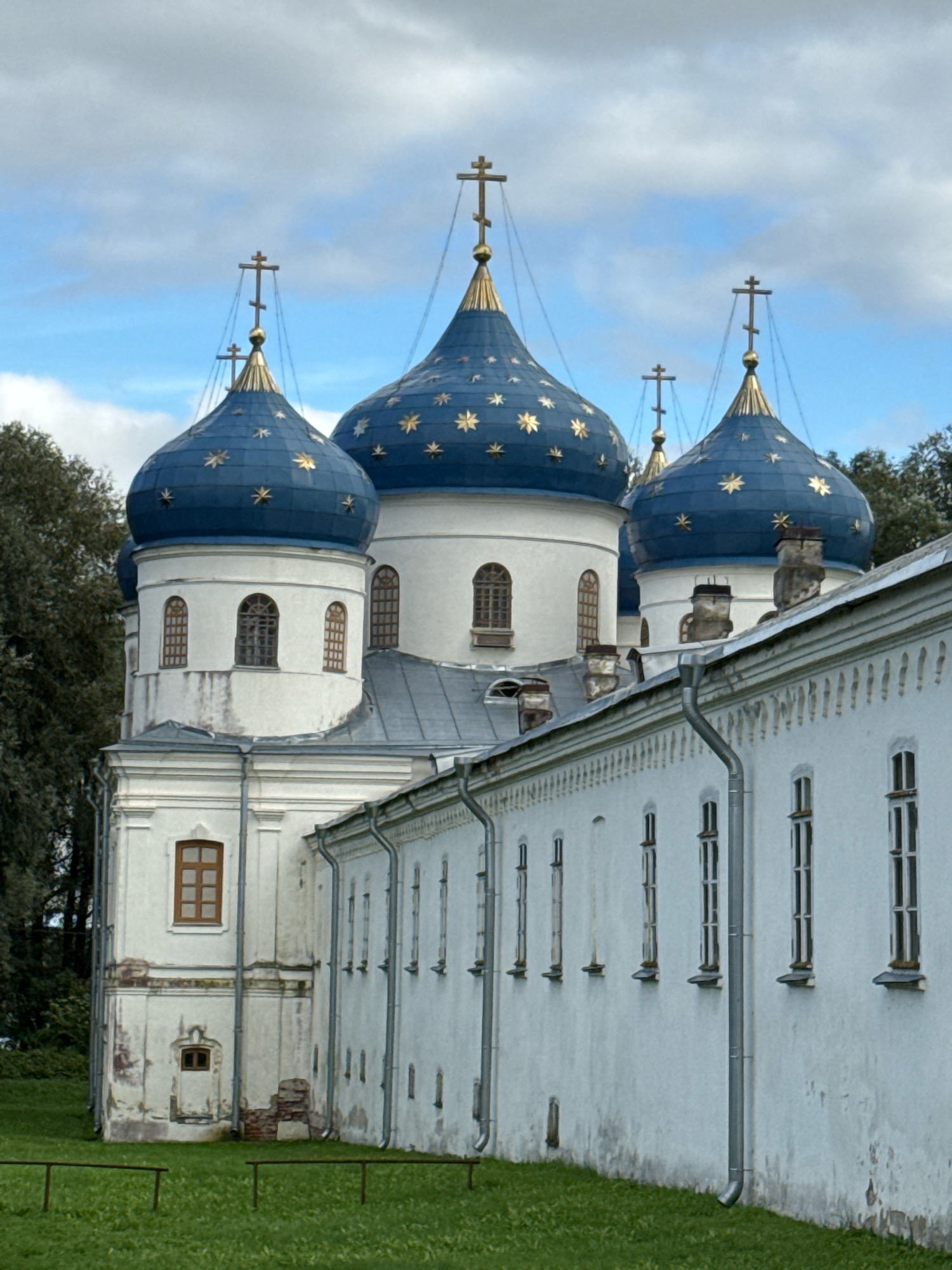 The image shows a view of the Saint Sophia Cathedral, a prominent landmark in Veliky Novgorod, Russia. The cathedral features several blue domes adorned with golden stars and topped with crosses. The white walls of the cathedral and an adjacent building are visible, with a row of windows along the side. Green grass covers the ground in front of the building, and trees are visible in the background. The sky is partly cloudy with patches of blue. There are no people or animals depicted in the image.