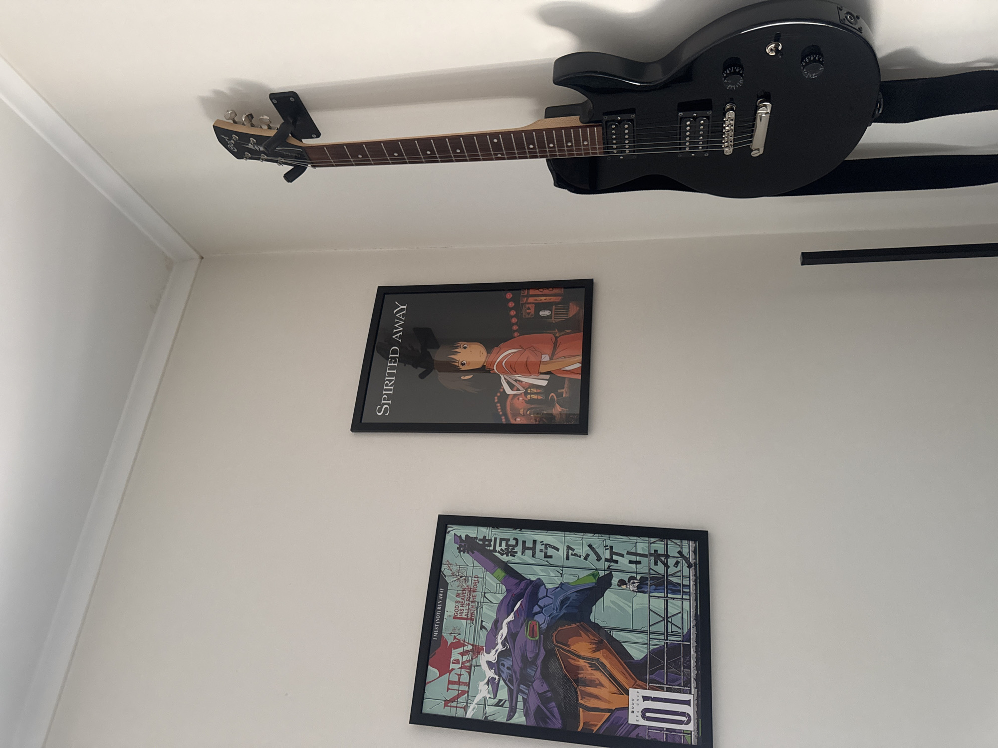 The image shows a corner of a room with two framed posters on a white wall. To the left, a poster features the character Evangelion Unit-01 from the anime series Neon Genesis Evangelion. To the right, a poster displays Chihiro Ogino from the Studio Ghibli film Spirited Away. A black electric guitar is mounted on the wall between the two posters, with its headstock pointing upwards. The room appears to be a living space or studio, with subtle architectural details such as crown molding visible at the top of the wall. The lighting is soft, casting gentle shadows. There are no people visible in the image. The scene is domestic and appears to be an interior setting. This image does not provide any information that could be used to determine its location in the city of Kremenchuk, Ukraine.
