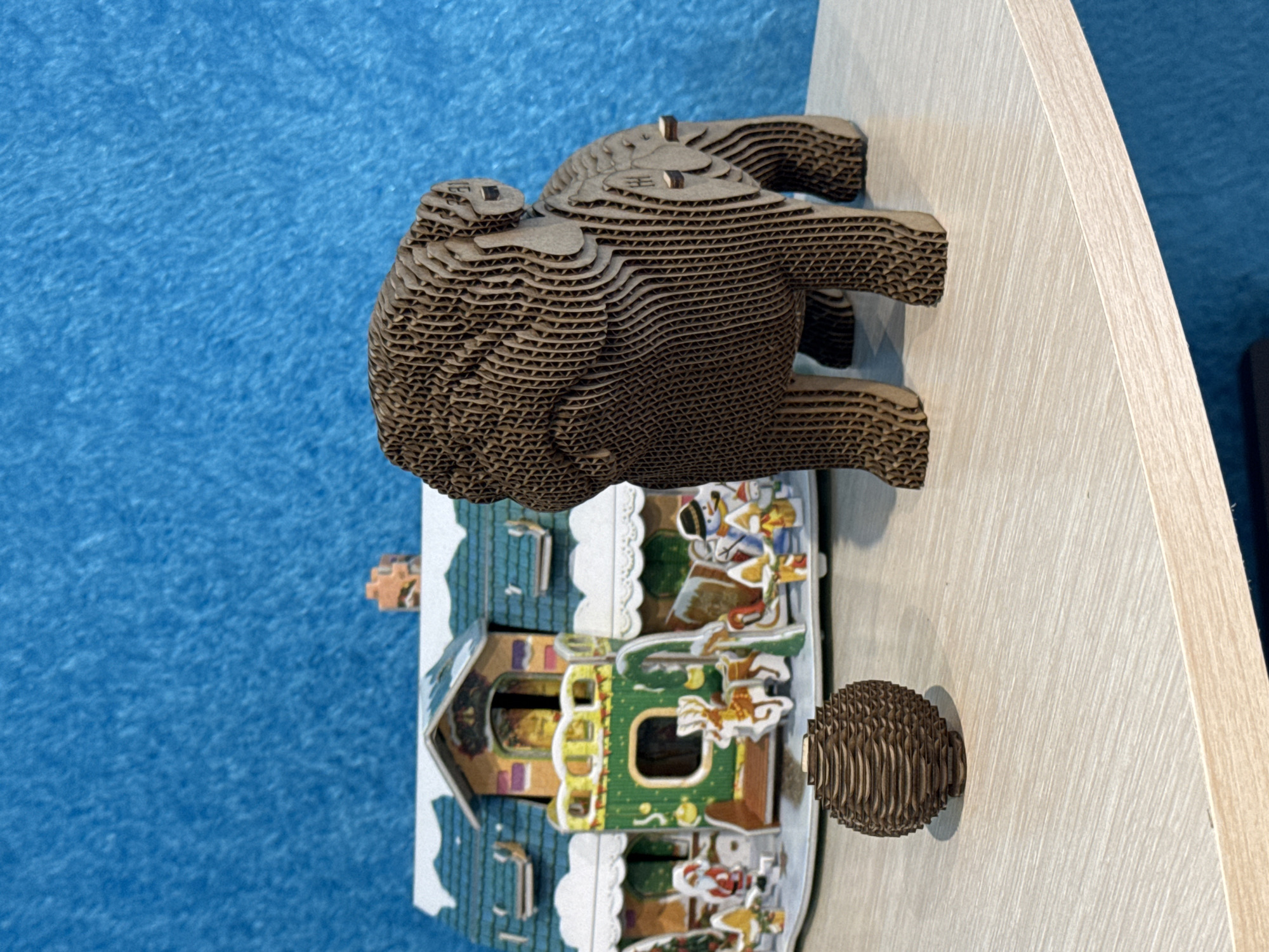 A cardboard Christmas-themed diorama sits on a light-colored wooden surface. The diorama depicts a house decorated for Christmas with cartoonish figures and green and yellow trim. Perched atop the house is a layered cardboard animal figure, possibly a bear, and to the right is a small, spherical cardboard object. The background consists of a blue, textured surface. The scene is in Chuprino, Russia.