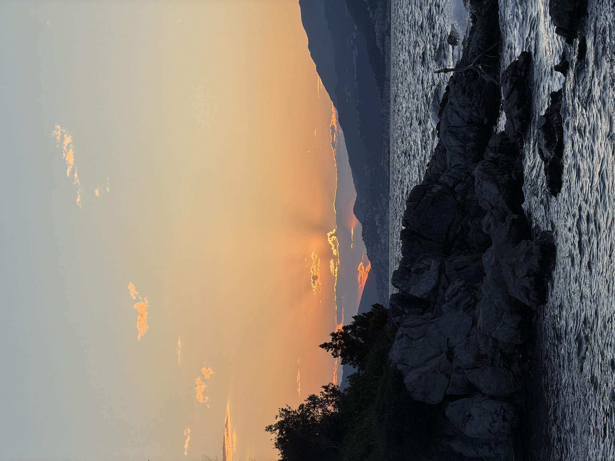 The image captures a sunset scene in Gošići, Montenegro. The sky is filled with orange and yellow hues, with light clouds scattered across the expanse. A silhouette of a mountain range is visible in the background. To the right, dark, rocky terrain and the water's edge are depicted. The water appears textured, possibly indicating movement or waves. The foreground features darker trees.