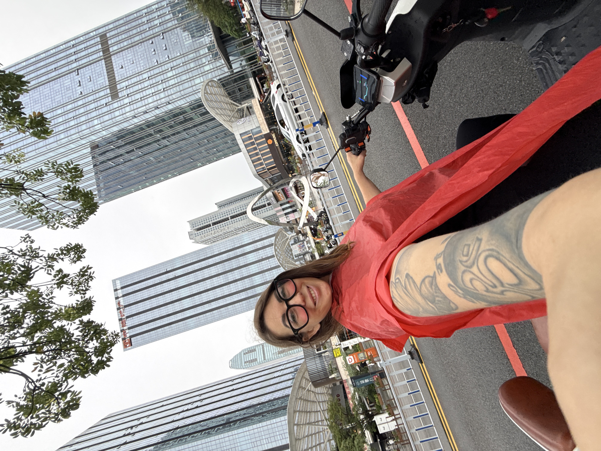 The image is a selfie taken from the perspective of someone sitting on an electric scooter in a modern urban environment. The person in the foreground is a woman with brown hair, wearing large black-rimmed glasses and a bright red rain poncho. She has visible tattoos on her left arm, which is resting on the handlebars of the scooter. The scooter's digital display is visible, showing a map or navigation interface.
In the background, towering glass skyscrapers dominate the skyline, characteristic of a major business district. One building prominently displays Chinese characters. The street is wet, suggesting it has recently rained. There are other vehicles, including cars and motorcycles, and pedestrians in the distance. An architectural structure resembling a large archway is visible on the left. The overall scene depicts a bustling city street in Huizhou, China, on a cloudy day.