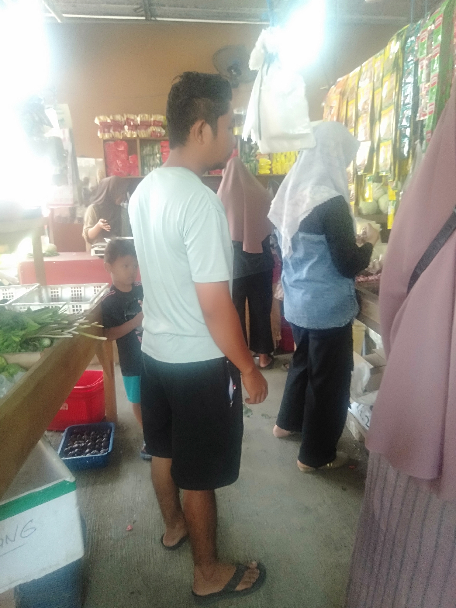The image depicts a bustling scene at an outdoor market in Sukadami, Indonesia. In the foreground, a man wearing a light blue t-shirt and black shorts stands with his back to the camera. To his left, a young boy in a black t-shirt and blue shorts is looking towards the left. Behind them, several individuals are browsing at market stalls. One woman wears a white hijab and a blue denim-like top, while another person in the background is partially visible. The stalls are laden with various goods, including what appears to be produce such as leafy vegetables and possibly fruits in a blue container. Bags of packaged items hang in the background. The ground is paved and appears to be a concrete surface. Sunlight streams in from the left, creating a bright, slightly overexposed area in the upper left quadrant of the image.