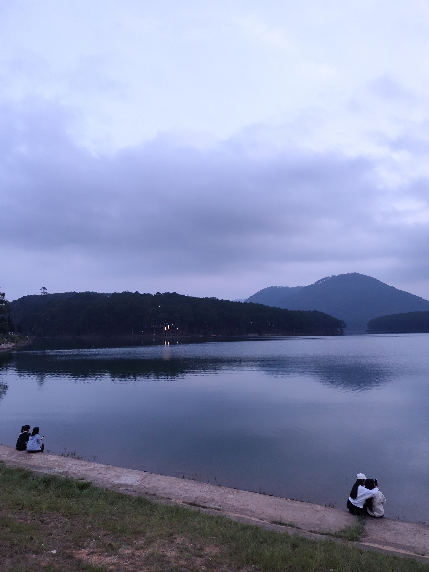 The image depicts a serene scene at Tuyen Lam Lake in Da Lat, Vietnam, under a cloudy sky. Two people are seated on a concrete embankment near the water's edge. Further down the embankment, another two people are sitting near the water. The calm lake surface reflects the sky and surrounding forested hills. Mountains are visible in the distance. The foreground consists of a grassy area leading up to the embankment.