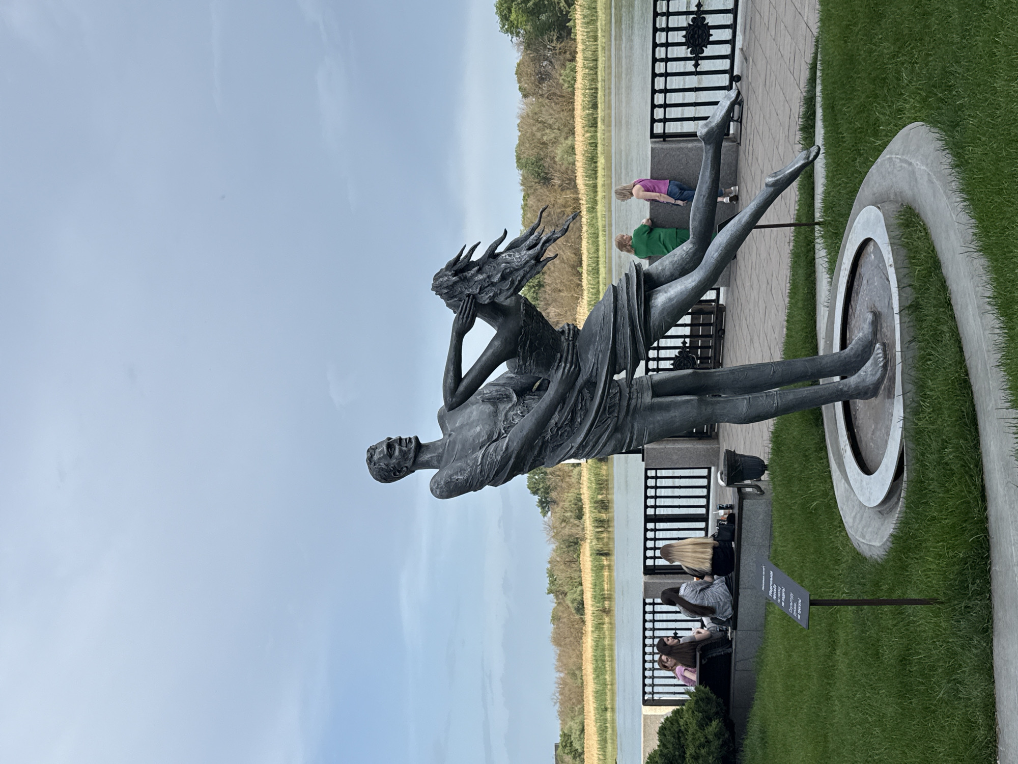 The image depicts a bronze statue of a man holding a woman in a pose that suggests a rescue or embrace. The statue is situated on a platform surrounded by a circular structure and grass. In the background, a body of water is visible, with trees and vegetation along its banks. A black metal fence runs alongside a paved walkway where people are sitting on benches. The location is Rostov-on-Don, Russia, as indicated by the language on the sign in front of the statue.