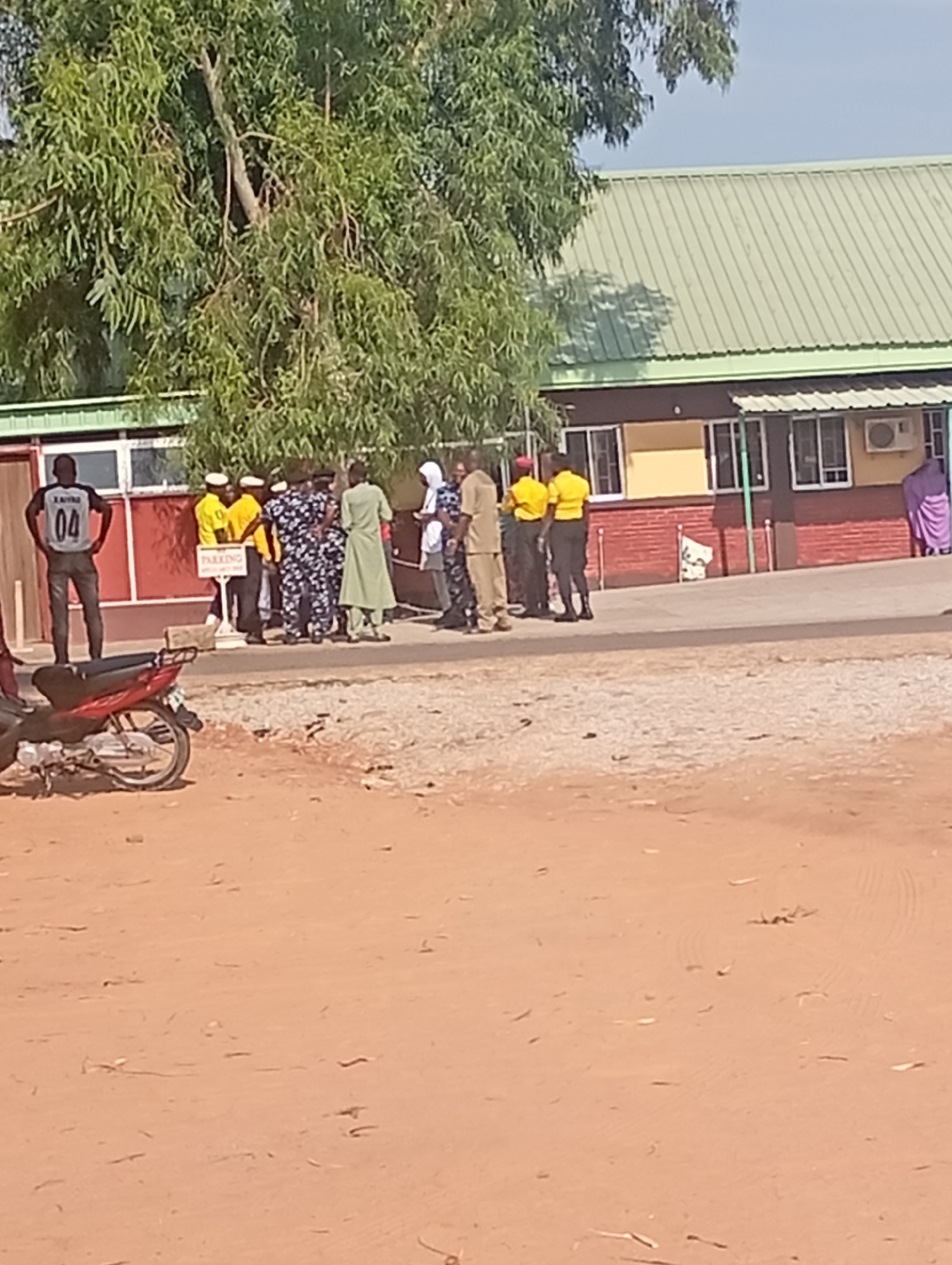 In Gombe, Nigeria, a group of individuals are gathered near a building with a green corrugated metal roof. A red motorcycle is parked in the foreground on a sandy surface. The building appears to be a single-story structure with multiple windows and an air conditioning unit visible on the exterior wall. A sign indicating "PARKING" is present among the group. The people are dressed in various attire, including uniforms and casual clothing, suggesting a mix of official and civilian presence. Some individuals are standing and talking, while others appear to be observing. The environment is outdoors with a tree providing shade.