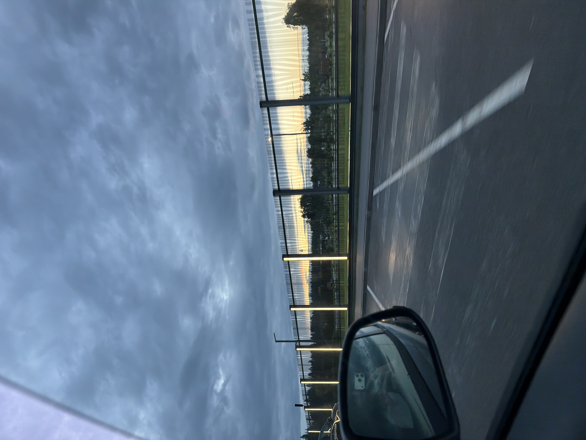 The image shows a cloudy sky over a cityscape in Solnechnoye, Russia. A reflective fence with vertical lights separates a road from trees and buildings in the background. A car's side mirror is visible in the foreground, reflecting a person and part of the car's interior. The road has white lane markings and appears to be a parking lot or an area with parking spaces. The lighting suggests it is either dawn or dusk.