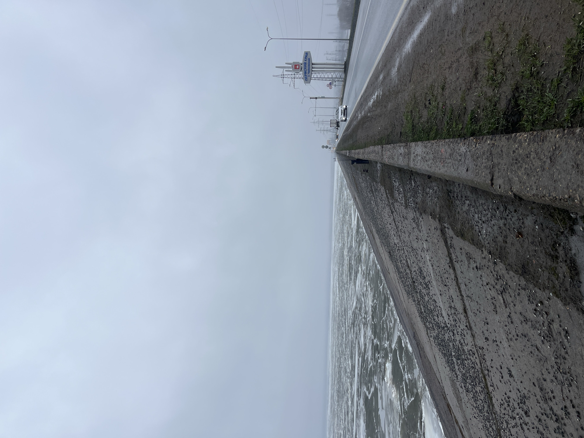The image depicts a bleak, overcast landscape featuring a wet road running alongside a large body of water, likely a river or reservoir. The perspective is from a low angle at the side of the road, looking forward.
The foreground shows a dark, weathered concrete barrier separating the road from the water. The water itself, occupying the left side of the frame, is covered with numerous chunks of ice and slush, suggesting cold weather or an early spring thaw. Above the concrete barrier, a muddy strip of ground with scattered green weeds leads up to the asphalt road. The road is dark and appears wet, possibly from rain or melting ice, with a faded white line marking its edge.
Along the right side of the road, stretching into the distance, are numerous utility poles, power lines, and streetlights. A prominent blue and white road sign is visible, displaying text in Cyrillic. The upper text reads "ПЕРМСКИЙ КРАЙ" (Permskiy Kray), identifying the region as Perm Krai. Below it, "г. Чайковский" (g. Chaykovskiy) is written, indicating the city of Chaykovsky. A small white car is visible on the road further down.
The sky is uniformly grey and overcast, contributing to the subdued and cold atmosphere of the scene. The image captures a daytime scene, though the heavy cloud cover prevents direct sunlight. No individuals are visible. The location cues on the sign directly place the scene in Perm Krai, near Chaykovsky, consistent with the provided location context of Noviy, Russia.