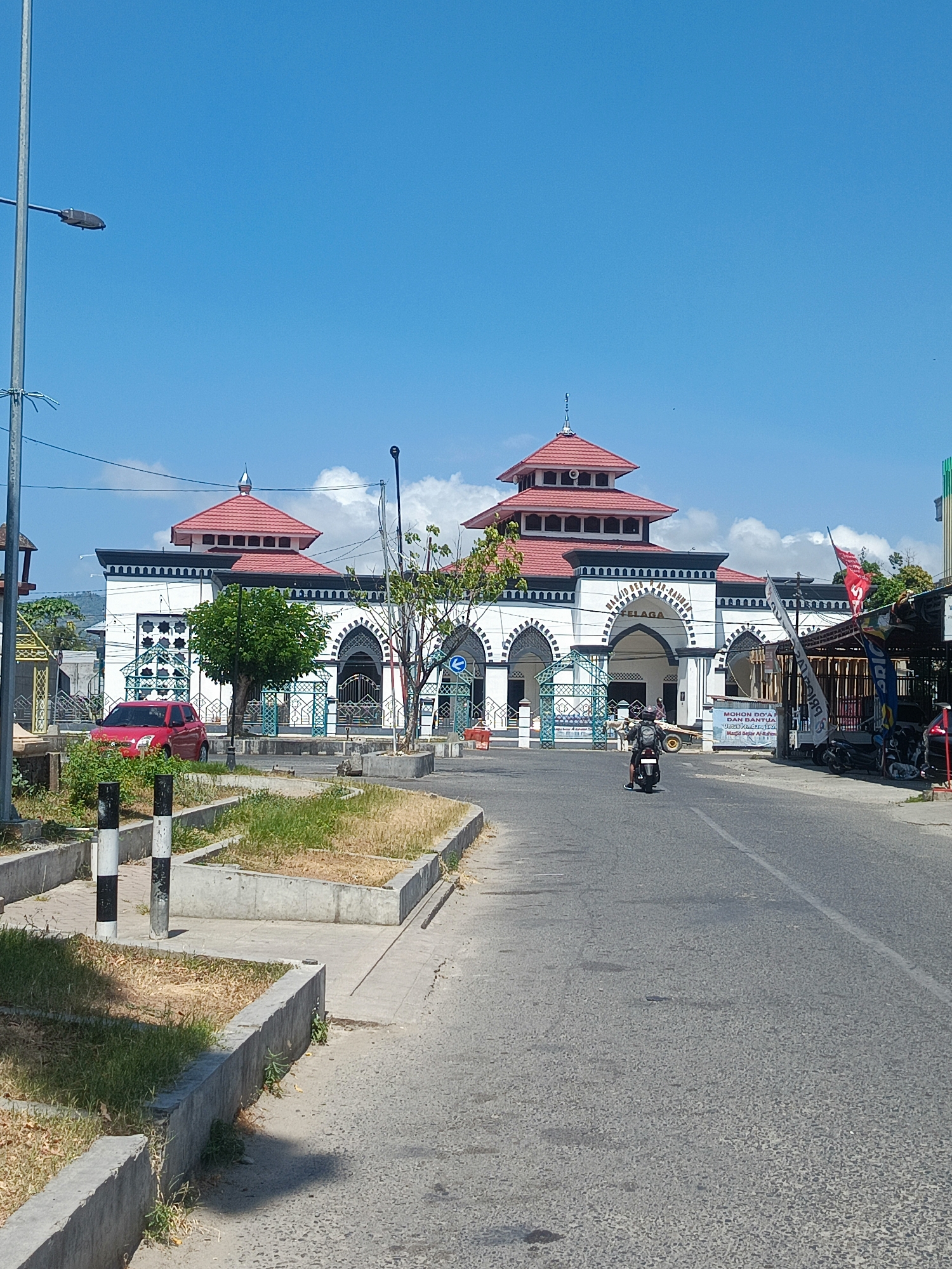 This is an eye-level, full shot image of a mosque in Luwuk, Indonesia. The mosque has white walls with black trim, and red tiered roofs. A white sign above the arched entrance reads "MASJID AR-RAHMAN LUWUK". In front of the mosque, there is a street with a single motorcycle rider in the foreground, heading away from the camera. To the left of the street, there is a red car parked next to a patch of dry grass and concrete planters. A street lamp is visible on the left side of the image. The sky is bright blue with scattered white clouds.