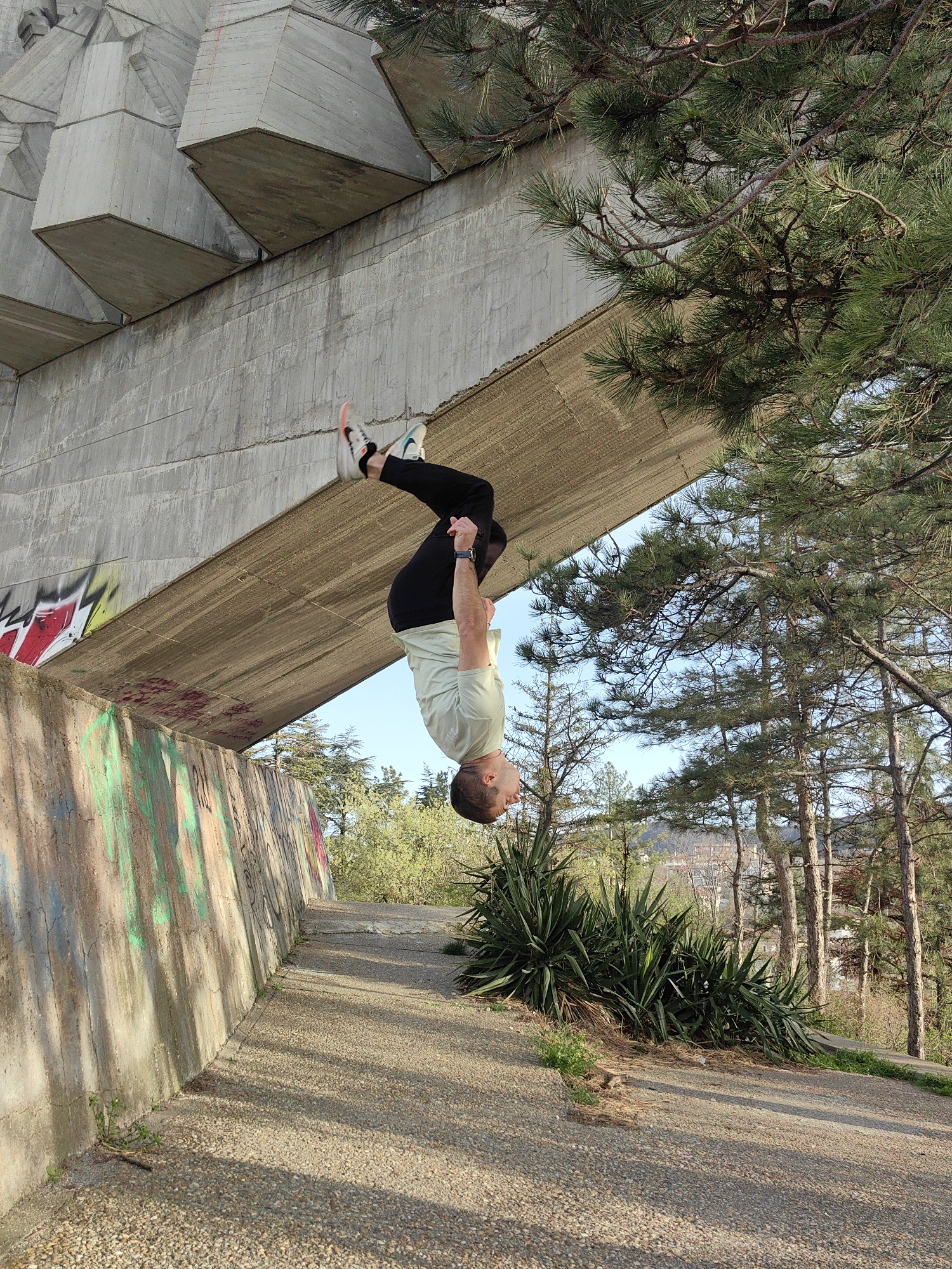 A man is upside down, hanging by his legs from a concrete structure. He wears black pants, a light-green t-shirt, and sneakers. The structure appears to be part of a bridge or architectural feature, with graffiti visible on the supporting wall. The background includes trees and foliage, suggesting a park or green space. The location is likely the Asparuhovo Bridge in Varna, Bulgaria, based on the architectural style of the concrete structure. The man is performing a physical feat of hanging upside down. The environment includes concrete structures, vegetation, and a paved path.
