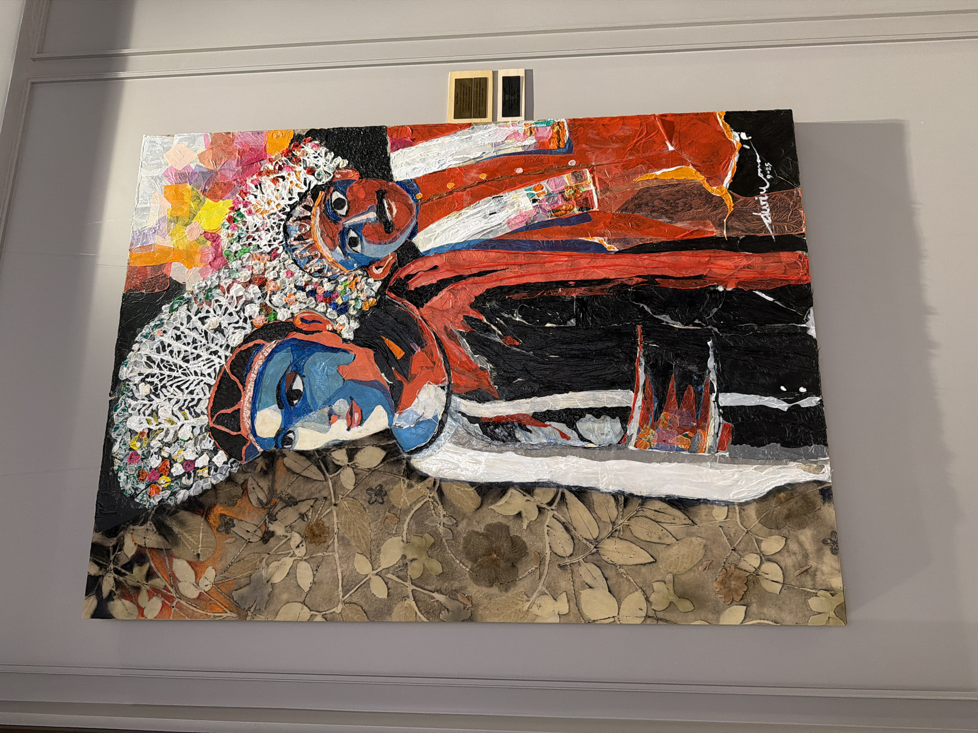 A painting hangs on a gray wall in an unknown location. The painting depicts two figures, one in black and red, the other in blue and white. The figures appear to be lying down. The bottom of the painting features a pattern of leaves. A small, dark object is mounted above the painting.