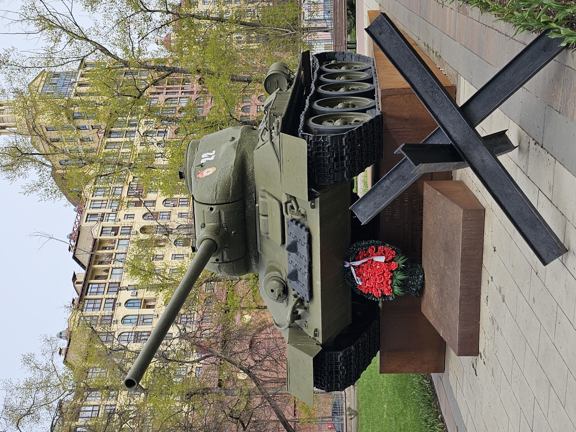 An olive green military tank, resembling a Soviet-era heavy tank, is prominently displayed outdoors as a monument in Khimki, Russia. The tank's long barrel points towards the left, and its tracks and road wheels are clearly visible. On the side of its turret, a white number "72" is painted, along with a red Soviet star emblem.
The tank rests on a reddish-brown, weathered metal or corten steel pedestal. Leaning against this pedestal, beneath the tank's front, is a wreath of vibrant red flowers adorned with a ribbon in the colors of the Russian flag (white, blue, and red). To the right of the tank, a black anti-tank obstacle, constructed from three intersecting I-beams, is positioned on the ground.
The pedestal's surface below the wreath bears engraved text, which includes the dates "1941-1945", signifying the Great Patriotic War (World War II), and fragments of words like "КРЫМ" (Crimea) and "СЕВАСТОПОЛЬ" (Sevastopol).
In the background, a tall, multi-story building with a light-colored facade, many windows, and some arched architectural details rises. Bare tree branches with emerging light green leaves partially obscure the building, suggesting springtime. The ground consists of paved walkways and patches of green grass. The sky is overcast, indicating daytime. The scene evokes a sense of solemn remembrance and historical commemoration.