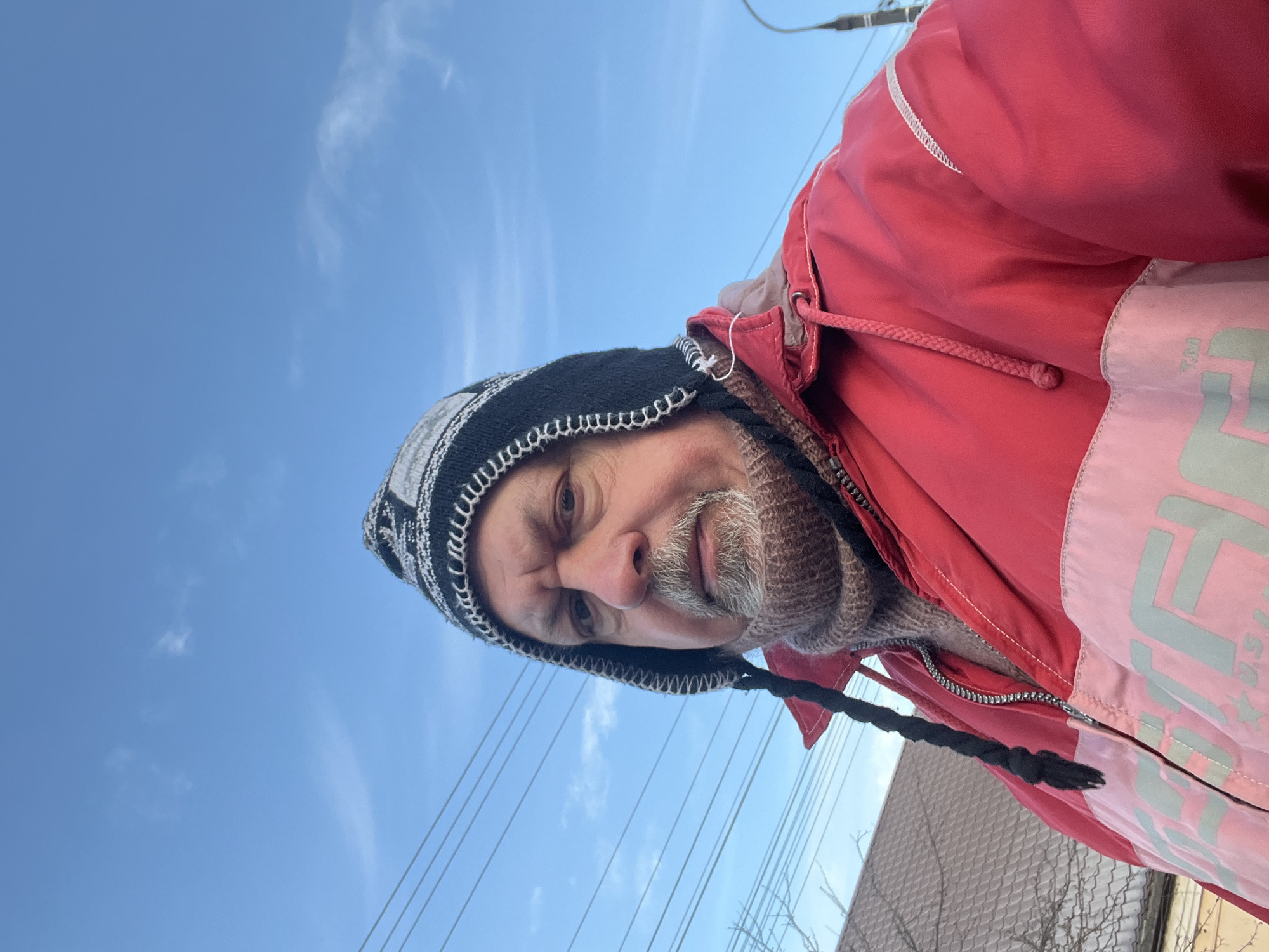 The image displays an adult male, observed from a low-angle selfie perspective, against a clear blue sky with sparse cirrus clouds. The subject, appearing middle-aged to older, has a gray beard and mustache. He wears a black, white, and gray knit winter hat with ear flaps, one of which features a black braided tie. A light brown or beige knit neck warmer is visible, covering his neck. His outerwear is a red and light pink two-toned winter jacket, featuring a prominent zipper. The light pink section of the jacket bears light gray or green lettering and a "T.M." symbol, possibly "USPA". The individual looks directly into the camera with a neutral expression.
The background elements are out of focus but reveal a residential or suburban setting. On the left, multiple horizontal utility lines are visible against the sky. Below these lines, a portion of a light-colored tiled roof is present, along with bare tree branches, suggesting a dormant season. On the far right, part of a dark pole, potentially a streetlight, is discernible. The specific location within the city of Podolsk, Russia, cannot be determined from the visual information presented.